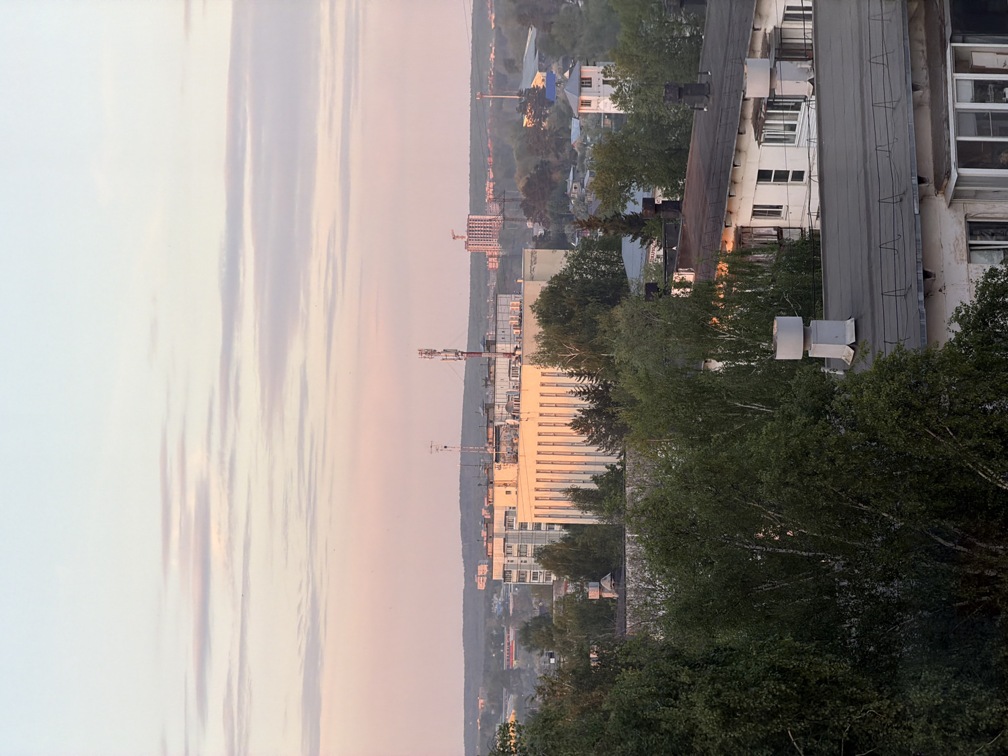 The image is a high-angle, daytime shot in Chaikovsky, Russia. A predominantly white high-rise building dominates the view, positioned amidst a scattering of lower buildings and dense, green trees. Communication towers are visible in the distance, rising above the city. To the left, a pale sky filled with streaks of light and clouds hints at either sunrise or sunset. A nearby apartment building with a grey roof and visible air conditioning units is also present, adding to the urban landscape. The scene is lit in soft, natural light, possibly during dawn or dusk.