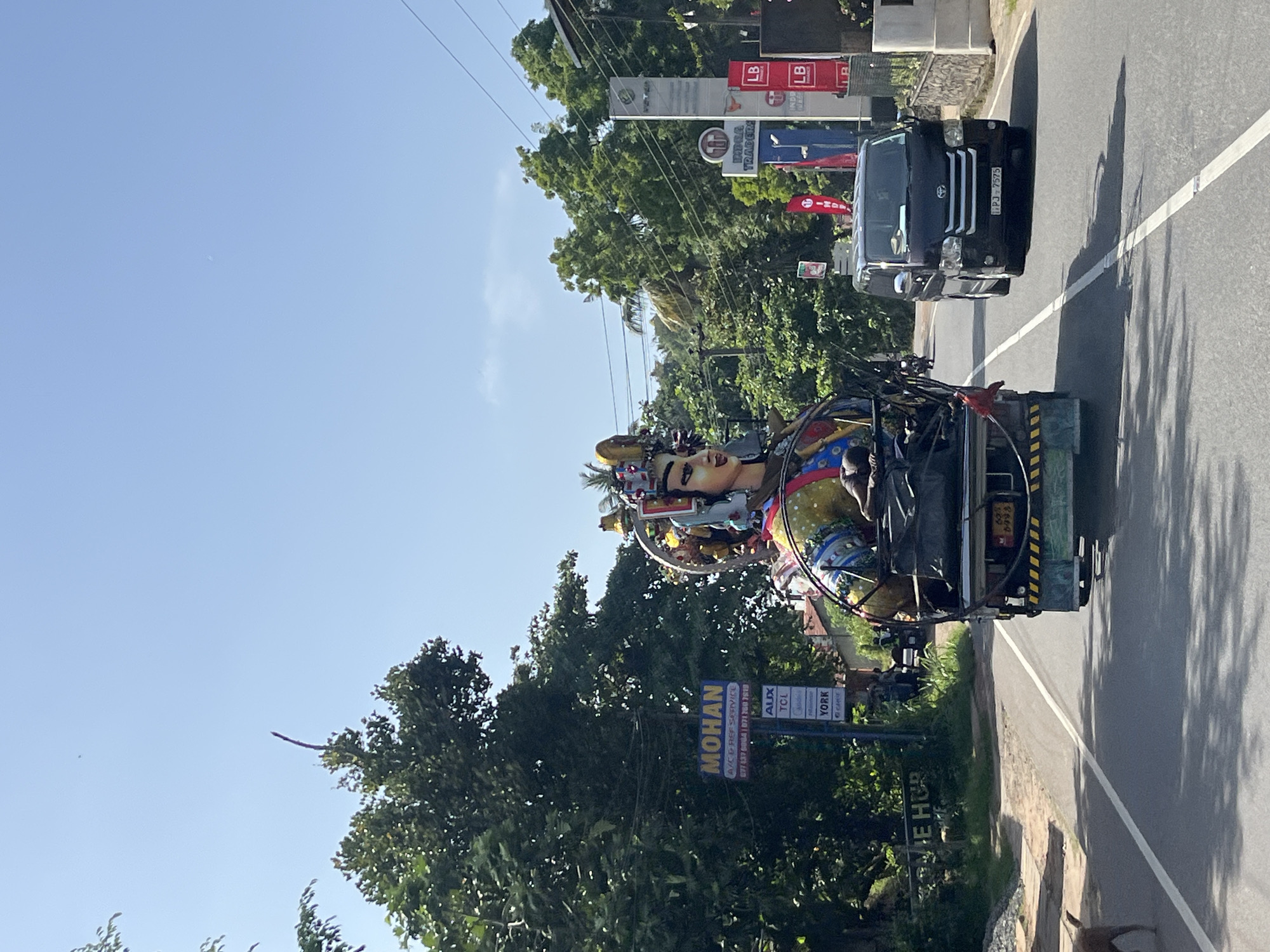 An eye-level, medium shot captures a vibrant, oversized effigy of a figure with exaggerated facial features being transported on the back of a truck. The effigy, adorned with a colorful headdress and detailed clothing, is secured to the truck's flatbed, which is piled with various items, including large yellow spheres and black netting. A man appears to be lying on top of the cargo, near the effigy's head. The truck is on a paved road with a white dashed line dividing the lanes, suggesting it's a typical street. In the foreground, a brown dog walks on the left side of the road. A dark-colored van travels in the lane next to the truck, its front license plate reading "PJ 7575". The roadside is lined with lush green trees and vegetation, indicating a tropical or subtropical environment. Several signs are visible, including one for "MOHAN A/C REF SERVICE" and another with "LB" logos. The sky is clear and blue. The image appears to be taken in Matara, Sri Lanka, due to the presence of the distinctive license plate format and the general visual cues of a Sri Lankan street scene.