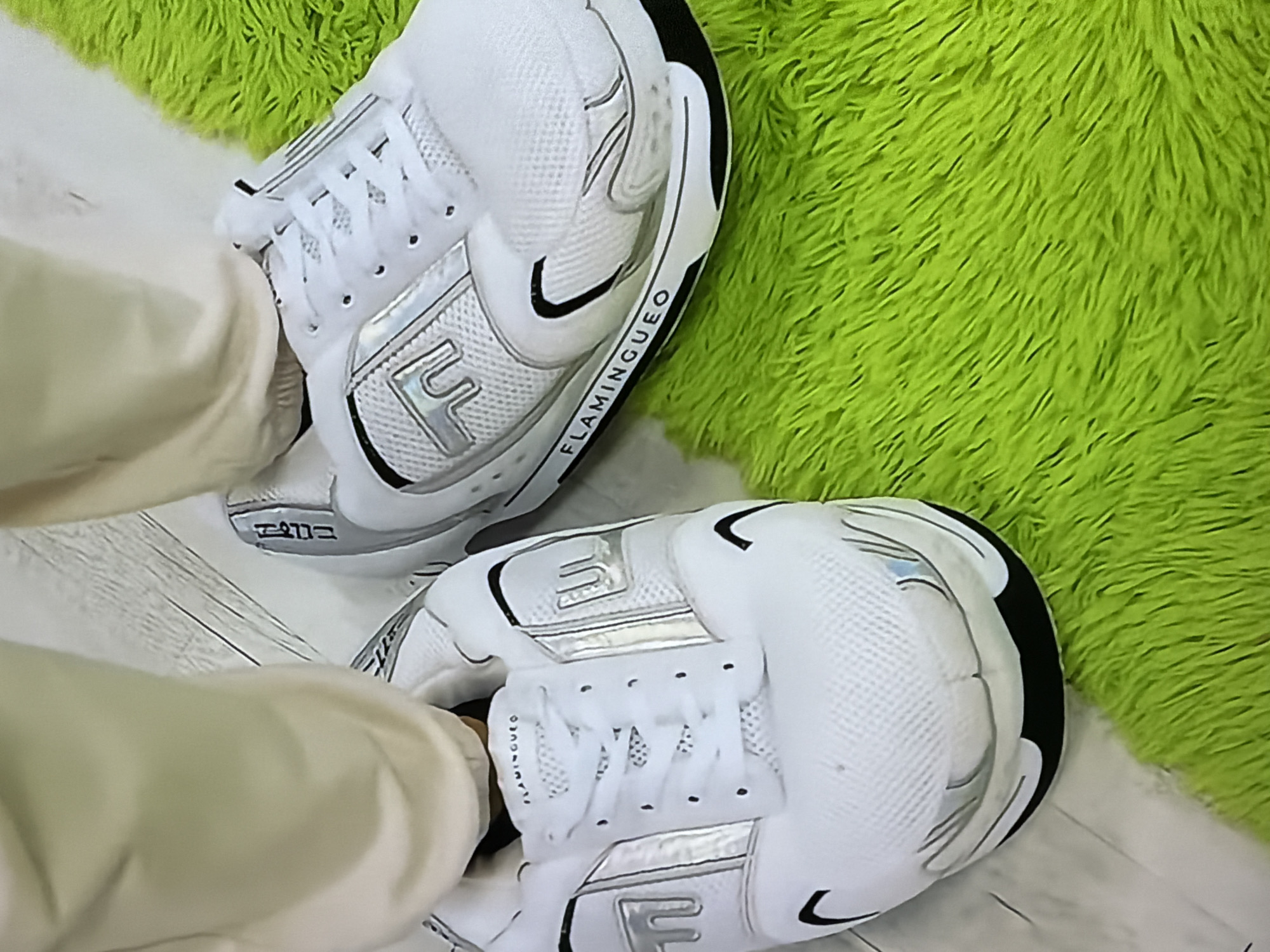 The image shows a close-up of two white sneakers with silver accents, resting on a vibrant green, fluffy rug. The sneakers appear to be casual athletic shoes, possibly house slippers designed to look like sneakers. One sneaker is partially obscured by light-colored pants. The word "FLAMINGUEO" is visible on the side of one of the sneakers. The background consists of a light-colored floor. There are no discernible elements in the image that would suggest a specific location in the city of Rome, Italy.