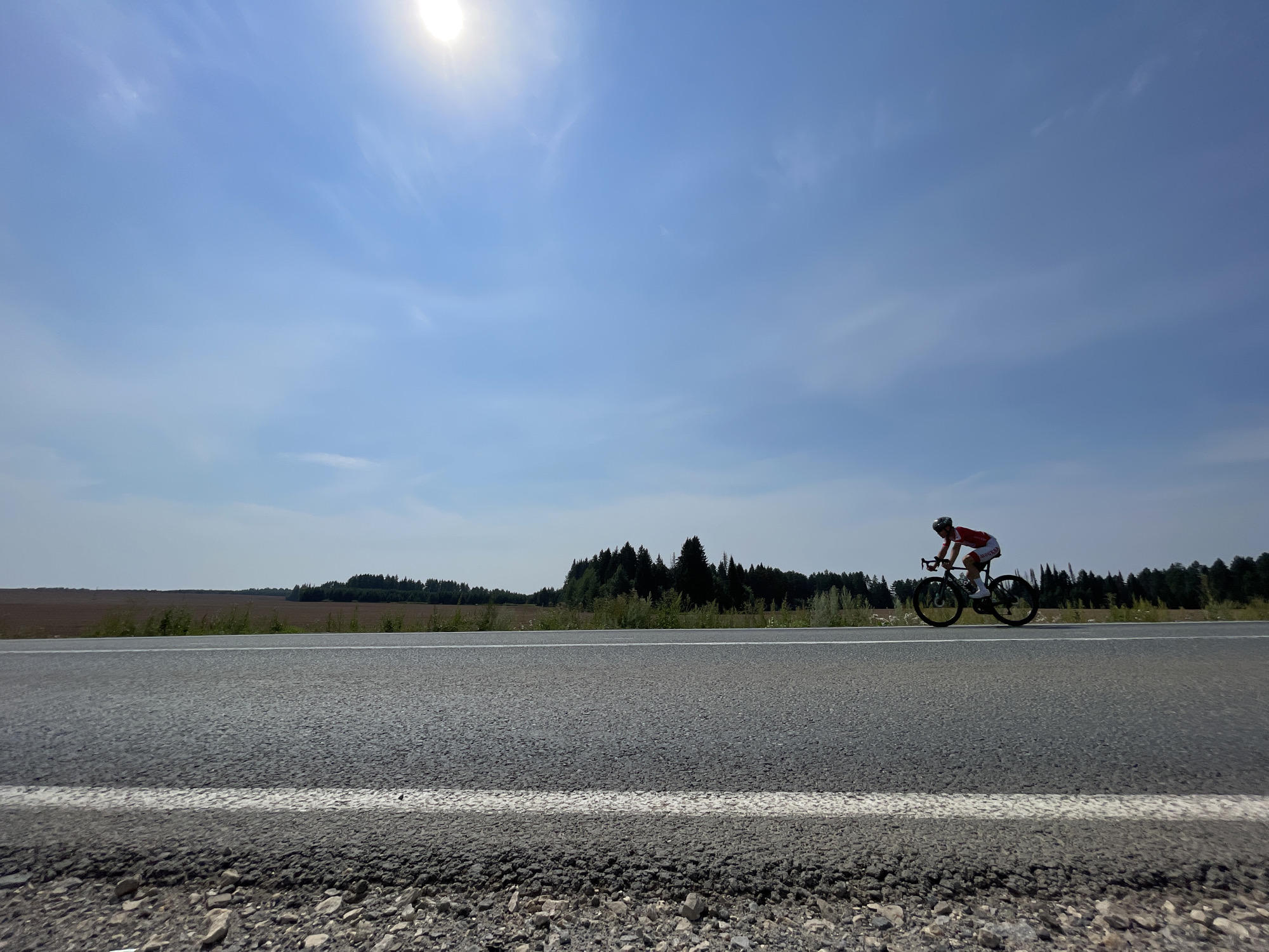 A person in a red and white cycling outfit rides a road bicycle along a paved road. The cyclist is wearing a helmet and appears to be in motion. The road has a white line marking its edge. In the background, a line of trees forms a dark band against a pale sky. The sun is visible in the upper left corner, partially obscured by wispy clouds. The foreground shows the rough texture of the road's shoulder, composed of small stones and gravel. The image depicts an outdoor scene under a clear, bright sky.