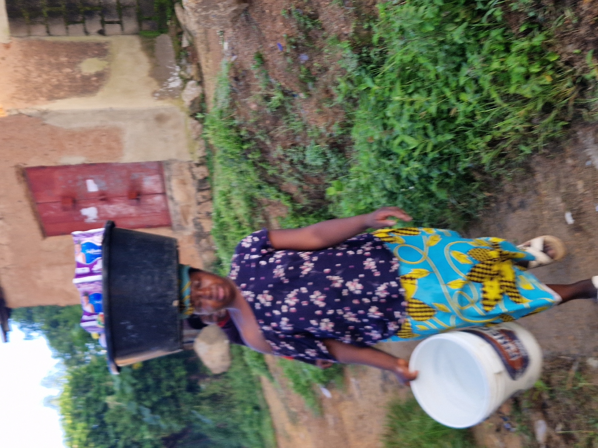 A woman carrying a large black basin filled with items on her head walks along a dirt path in front of a rustic, tan-colored building with a red door. She is also holding a white bucket in her right hand. She is wearing a dark blue shirt with a floral pattern and a vibrant blue and yellow patterned skirt. Another person is partially visible behind her. The surroundings are characterized by lush green vegetation and uneven terrain. The overall scene depicts a daily activity in a rural or semi-rural setting.