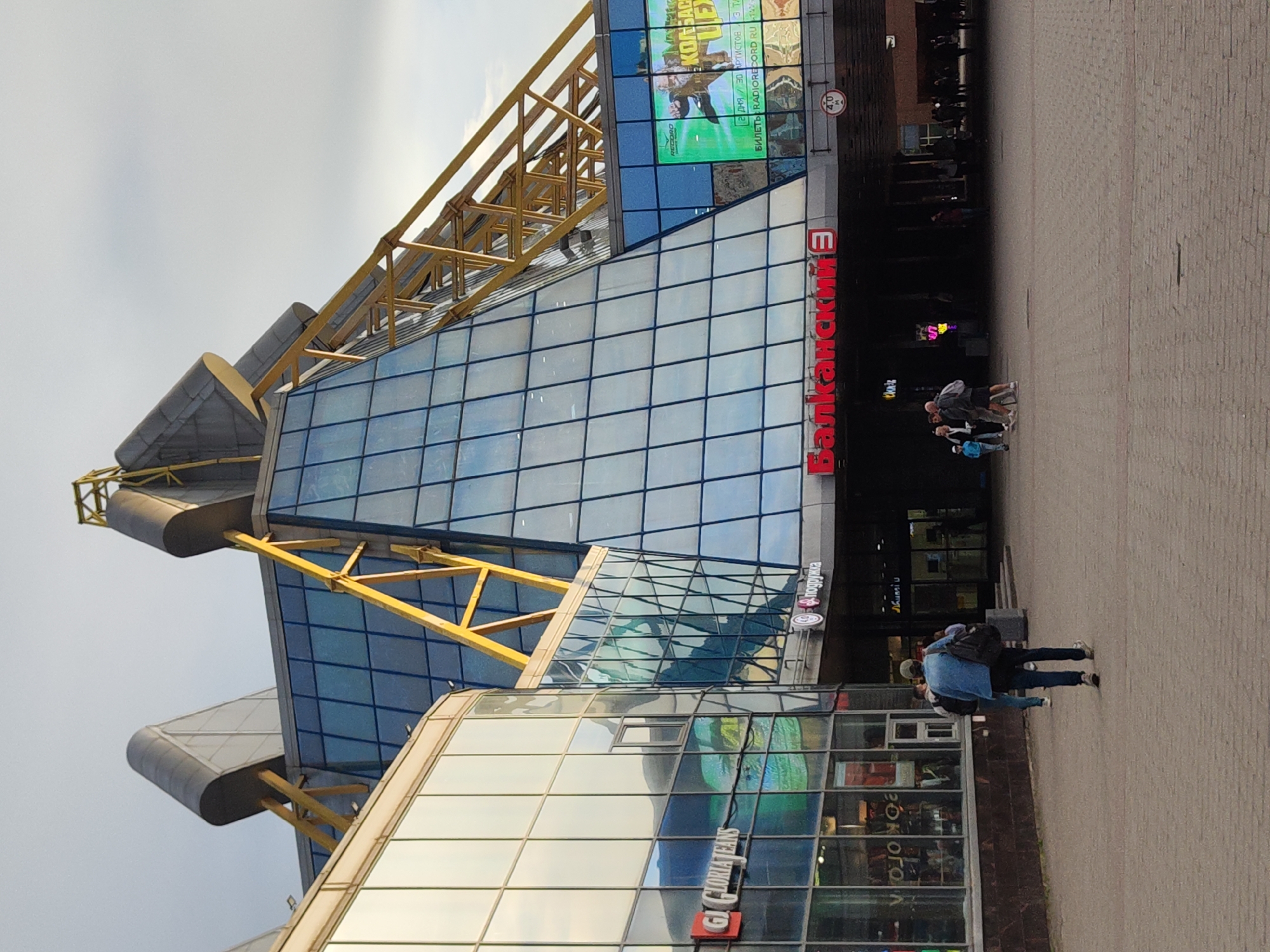This is a medium shot photograph taken from a low angle, looking up at a modern building with a distinctive architectural design. The building features a combination of blue-tinted glass panels and yellow structural elements that create a striking visual contrast. The sky is overcast and a dull grey, suggesting cloudy or perhaps late afternoon weather.
In the foreground, a paved plaza area is visible, with several people walking. On the right side of the image, a group of three people are walking away from the camera, their backs to the viewer. One individual is wearing a blue jacket and dark pants, another a dark jacket and jeans, and the third a light-colored shirt and dark pants. They appear to be casually strolling. Further back, other figures are visible walking into the building's entrance.
The building itself has a large glass facade with a grid pattern of frames. There are also sections of metallic grey and yellow structural supports. A prominent red sign on the building reads "Балканский" (Balkansky) in Cyrillic letters, suggesting this is a shopping center or a commercial building. Above this, a large digital screen displays colorful imagery, possibly an advertisement or event promotion. Text on the screen and on smaller signs is visible, including "RECORO," "KONSAC," and "GLORIATEANS," as well as event details like "2 дня 30 АРТИСТОВ" (2 days 30 artists). There is also a traffic sign indicating "4.0" possibly a height restriction. The overall impression is a bustling urban environment with modern architecture.