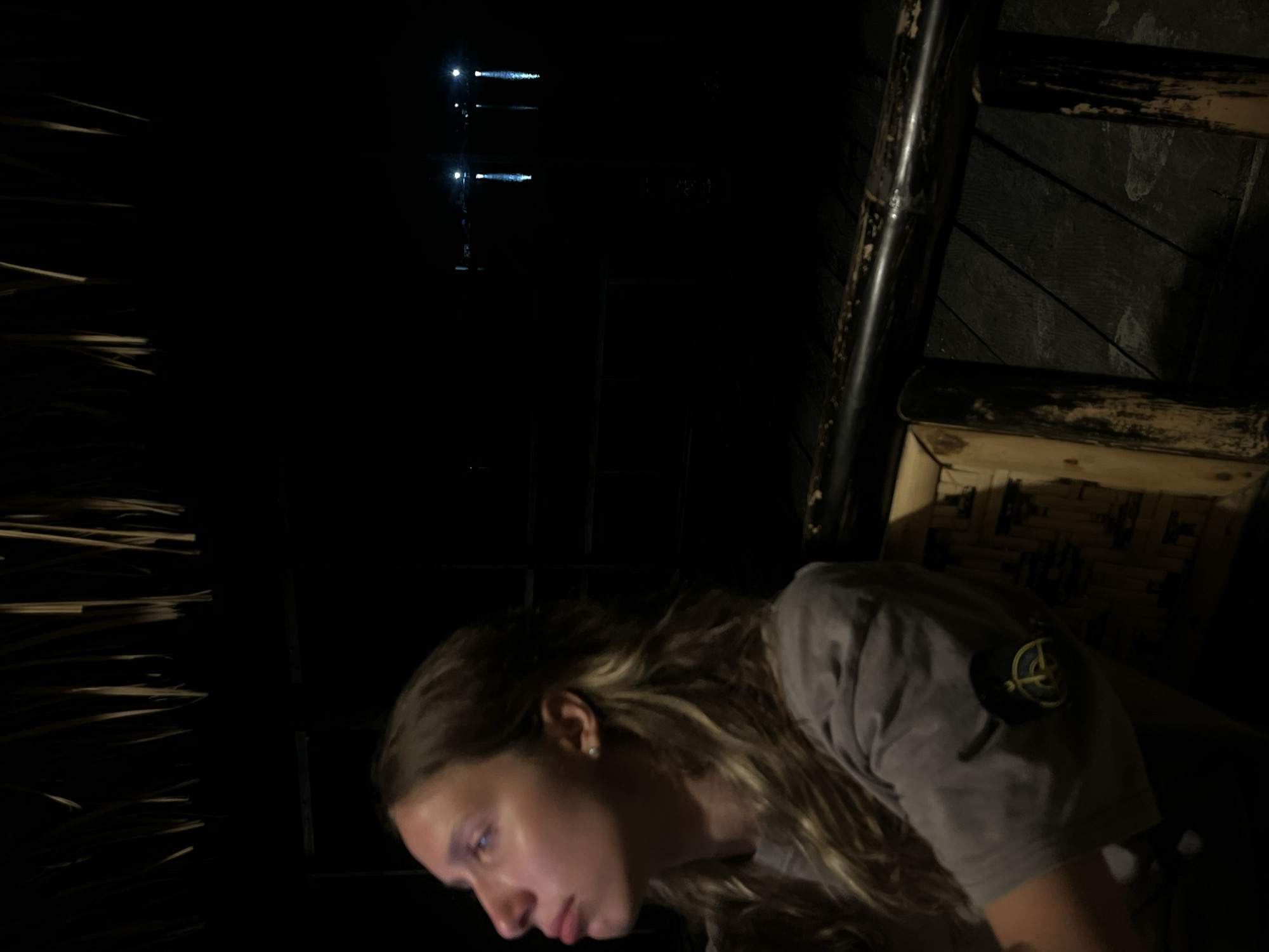 A young woman with light brown hair, wearing a grey t-shirt with a circular patch on the sleeve, is seen from the chest up, looking upwards to her left. She is sitting on a bamboo chair in what appears to be an outdoor dining area. The background is dark, with hints of natural thatch overhead and a dimly lit body of water with lights reflected on it in the distance. The scene suggests a nighttime setting, possibly at a restaurant or resort in Kertasari, Indonesia, a location known for its coastal views and natural beauty.