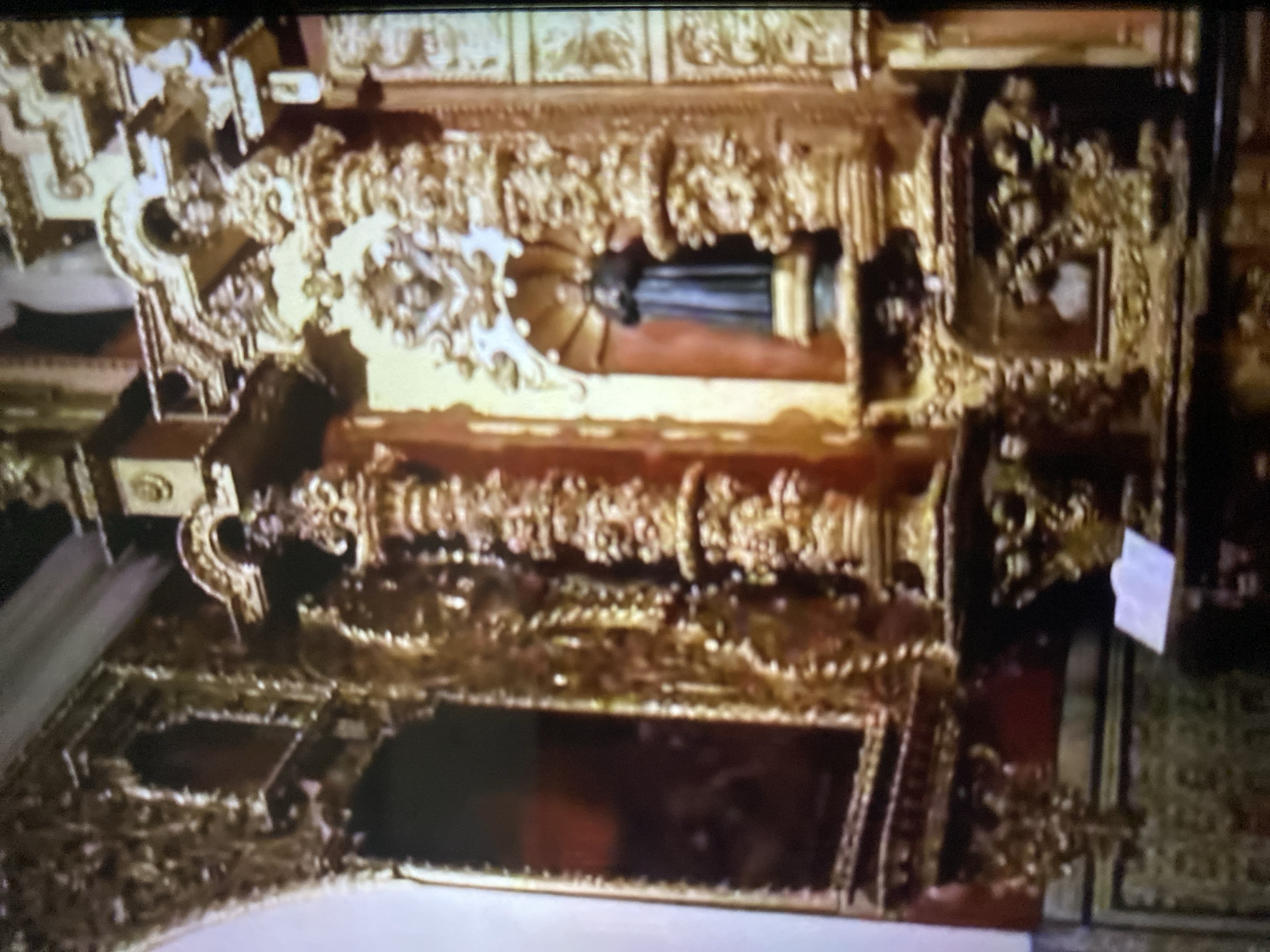 A low-angle shot depicts an ornately gilded altar inside a church or cathedral in Tinaquillo, Venezuela. The altar is multi-tiered and densely decorated with carvings and sculptural details. A dark-colored statue is positioned in a central niche on an upper tier. Below, other details include carved columns, scrolls, and possibly religious iconography. A paper or tag is visible on the right side. The overall environment is rich and historical.