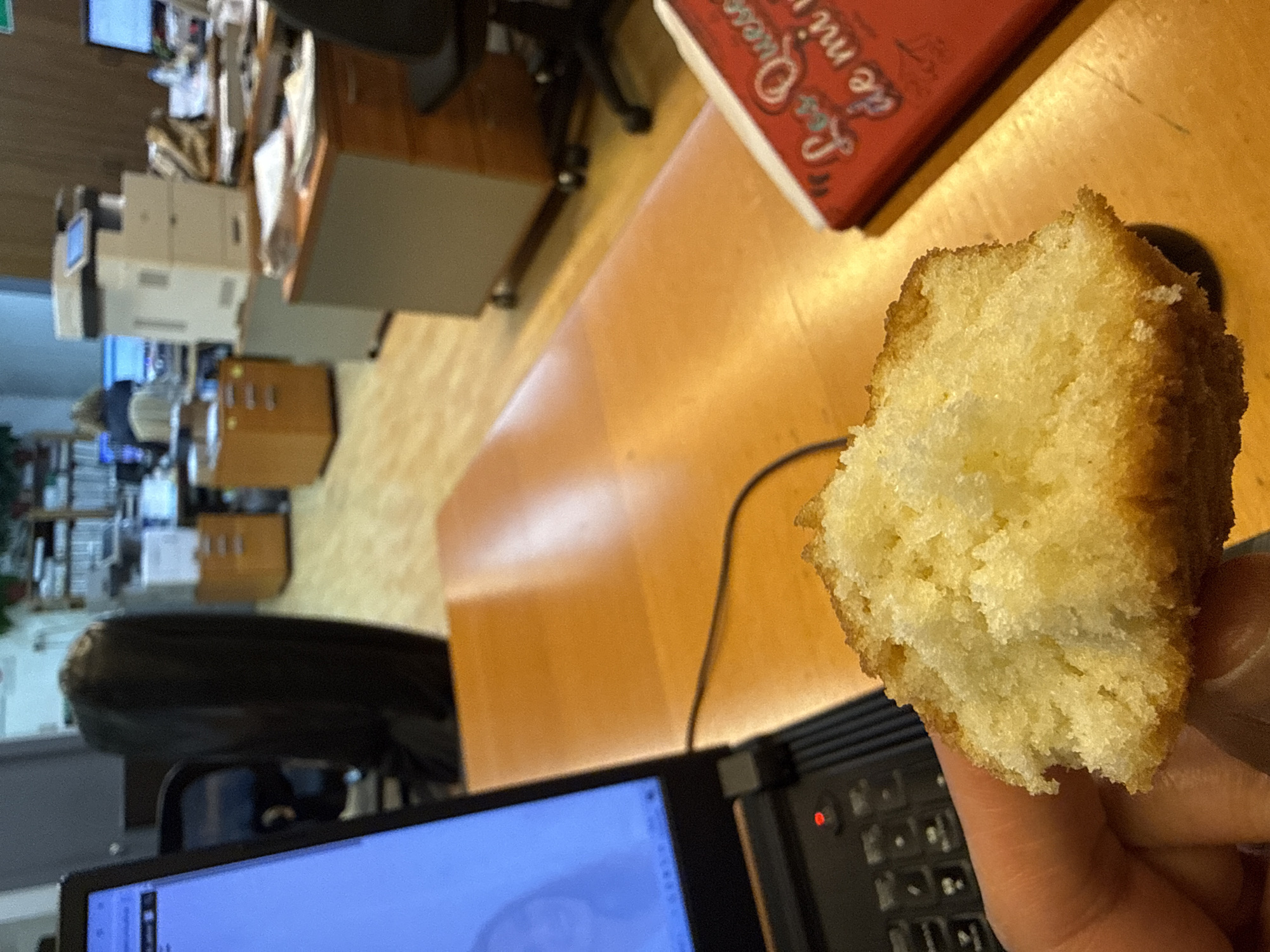 The image depicts a partially eaten piece of cake held in a person's hand. The background shows an office environment with desks, chairs, computers, a printer, and file cabinets. A book with the title "Les Quesse" in red lettering is visible on the desk. The exact location within Barcelona, Spain is impossible to determine from the image alone.