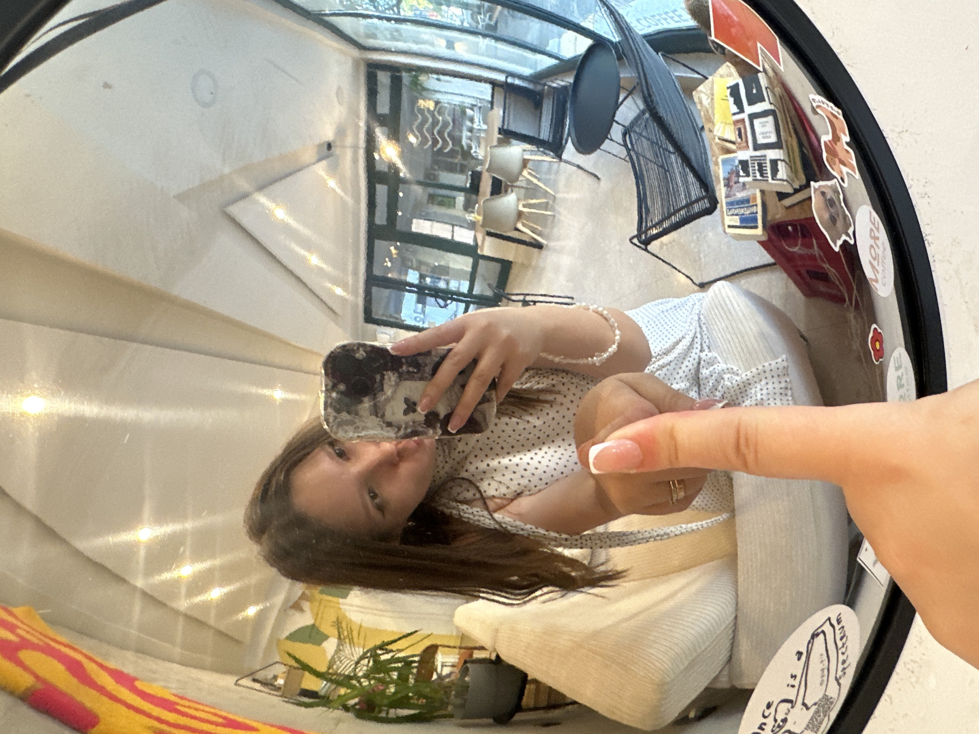 The image shows a person reflected in a round mirror with a black frame, which is adorned with various stickers and items. The person in the reflection is holding a smartphone and taking a selfie. The person's face is partially obscured by the phone, but their long, brown hair is visible. They are wearing a white shirt with black polka dots and a pearl bracelet. The fingertip of another person extends into the frame. The background reflected in the mirror shows what appears to be the interior of a cafe or restaurant, with tables, chairs, a skylight, and decorative elements. Based on the style of the cafe, this photo may have been taken in Budva, Montenegro, but there is not enough definitive information to be certain.