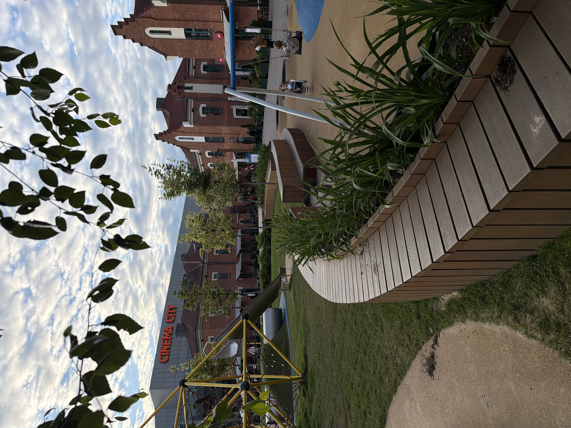 An outdoor playground area is depicted, with a modern climbing structure featuring a yellow frame and a gray slide on the left. Adjacent to it, a curved wooden bench with planters filled with green foliage runs along the foreground. Beyond the playground, there are red brick buildings with arched windows and gabled roofs, reminiscent of historical architecture. A large, contemporary building with the illuminated text "CINEMA CITY" is visible in the background. In the middle ground, children are playing on swings and a spring rider. The ground is a mixture of grass, sand, and colored rubber surfacing. The sky is partly cloudy. This scene is located in Krakow, Poland, specifically at thearyan Entertainment Center, near the historic Jewish Quarter.