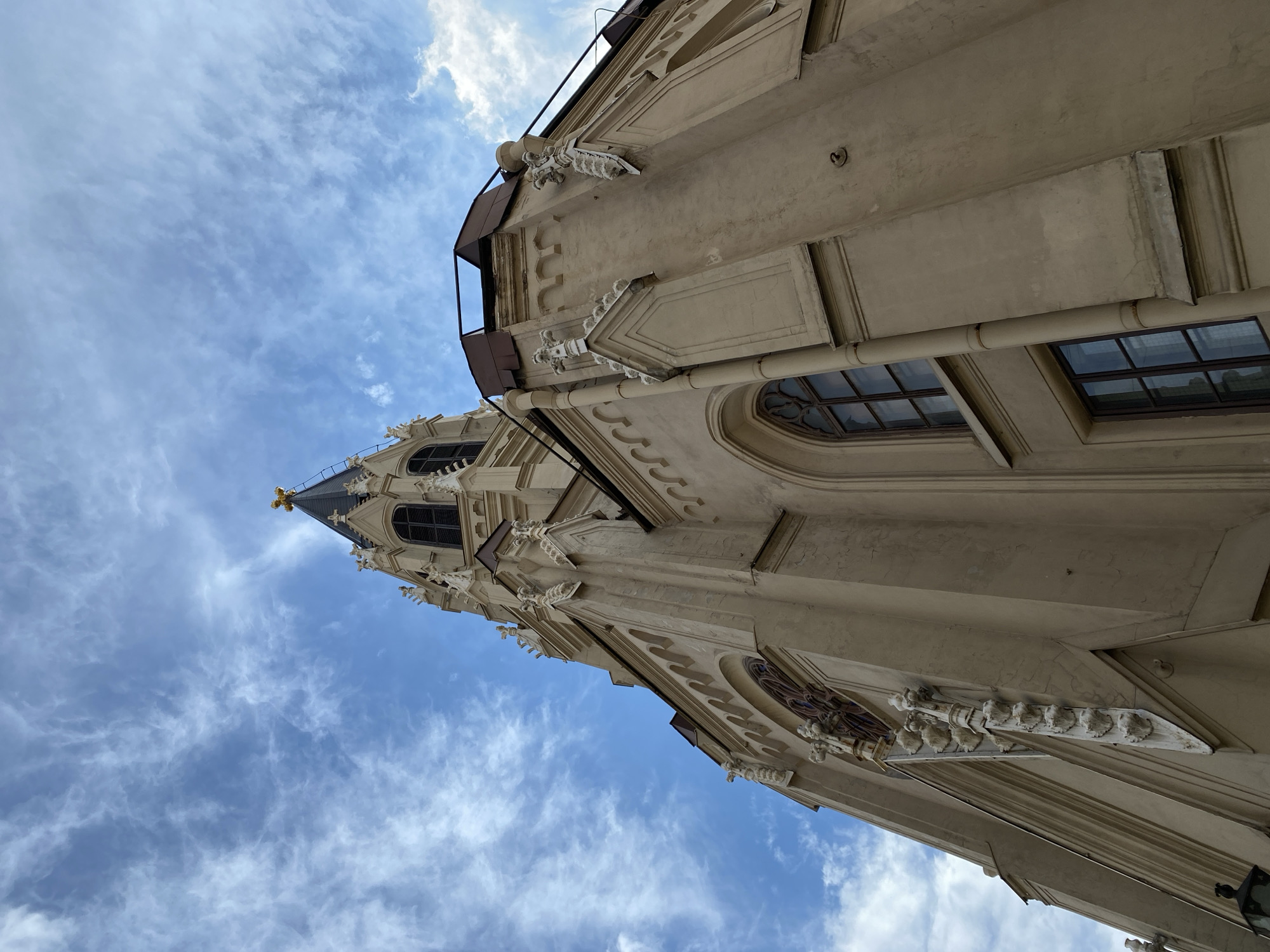 This is a low-angle, wide-shot photograph of the Church of the Assumption of the Blessed Virgin Mary (also known as the Catholic Church of the Assumption) in St. Petersburg, Russia. The facade of the church is a light beige color with Gothic architectural elements. A tall, pointed spire dominates the upper portion of the frame, topped with a golden cross. The sky is a vivid blue with wispy white clouds scattered across it. Several arched windows with dark frames are visible on the side of the church, and ornate decorative elements are present along the roofline and on the walls. The perspective emphasizes the height and grandeur of the building against the expansive sky.
