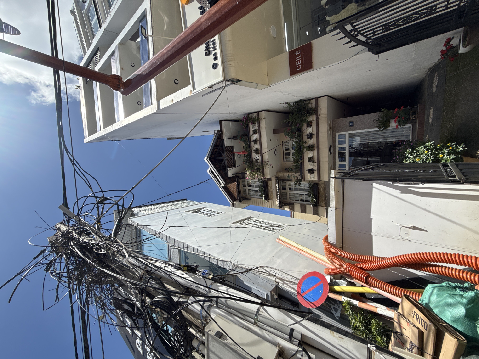 The image shows a narrow street in Da Lat, Vietnam, with several buildings packed closely together. A tangled mess of electrical wires dominates the left side of the frame, connected to a concrete pole. To the right of the wires, a narrow, light-colored building stands tall. Further to the right, a white building with multiple balconies and windows is visible. In the foreground, on the left, a red and blue "no parking" sign is mounted on a pole, alongside orange corrugated pipes and stacked cardboard boxes covered with a green tarp. The street itself is paved with stone slabs. The buildings display architectural elements characteristic of Da Lat, including sloping roofs and balconies adorned with plants. The overall impression is one of dense urban development with a mix of residential and commercial spaces, characterized by a noticeable degree of urban clutter, particularly in the form of overhead wiring.