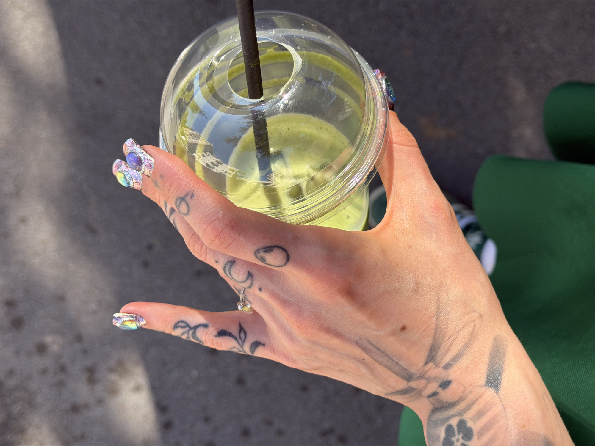 A person's hand holds a clear plastic cup containing a green smoothie with a black straw. The hand features multiple tattoos and ornate nail art with iridescent gemstones. A small ring adorns a finger. The background consists of a grey surface and a green object, potentially a chair or bench. Based on the appearance of the cup and the surrounding environment, the image was likely taken outdoors. The location is not able to be determined based on the image.