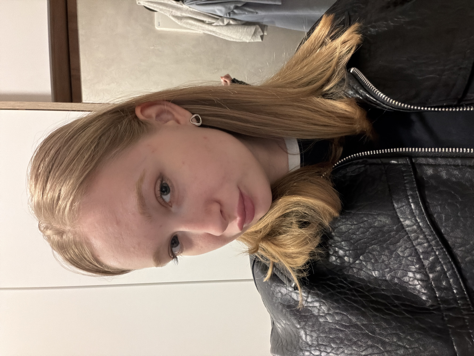 A young Caucasian woman with blonde hair is facing the camera, wearing a black leather jacket. She has fair skin and a stud earring. The background includes a light-colored wall, a white door, and a garment rack with clothes. The location is unknown, as there is nothing specific to indicate it is in Rossiyskiy, Russia.