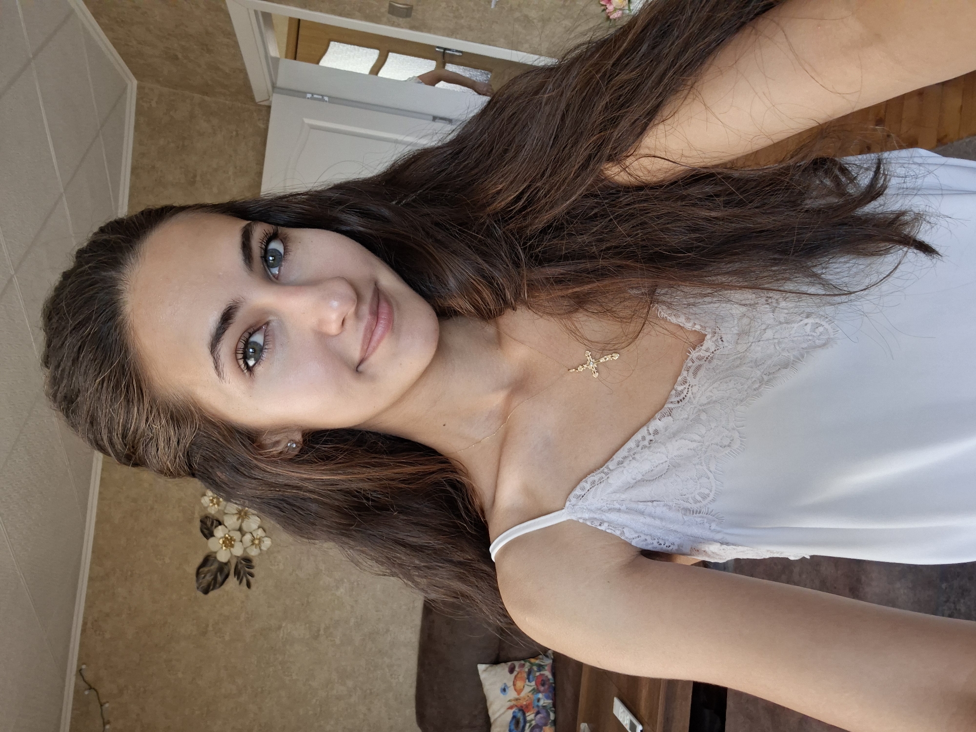 In a well-lit indoor setting, a young woman with long, dark brown hair and striking blue eyes poses for a selfie. She is wearing a white lace-trimmed camisole and a gold cross necklace. The background reveals a room with textured beige wallpaper, a brown sofa with a colorful pillow, a wooden table with a remote control, and a partially open door. The overall scene suggests a casual, domestic environment.