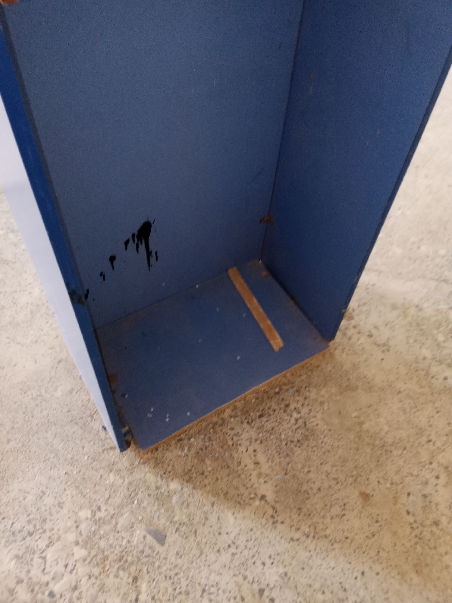 This is a close-up, eye-level, medium shot of the interior of a dark blue, rectangular object, possibly a cabinet or bin, on a concrete floor. The object is open at the top and appears to be made of a hard material like wood or plastic. The interior is predominantly a matte dark blue color.
On the back wall of the interior, there is a significant dark, amorphous stain, likely paint or a liquid spill, with several smaller splatters radiating from it. The stain is roughly in the center of the visible back wall.
The floor of the interior is also dark blue and appears to be slightly worn, with some lighter-colored specks visible. There is a single horizontal piece of light brown material, possibly wood or cardboard, adhered to the floor near the front edge of the interior.
The exterior of the object is partially visible on the left side, revealing a white surface with a blue edge. The exterior surfaces of the blue object show some signs of wear, with small, lighter-colored flecks scattered across them.
The surrounding environment, visible in the background and on the floor, is a textured, light brown or tan concrete surface with various dark and light colored aggregate. There is a shadow cast from the object onto the floor, indicating an overhead light source. The scene is outdoors or in a well-lit indoor space with a utilitarian atmosphere. The presence of Jalingo, Nigeria, as a location is not discernible from the visual evidence in the image.