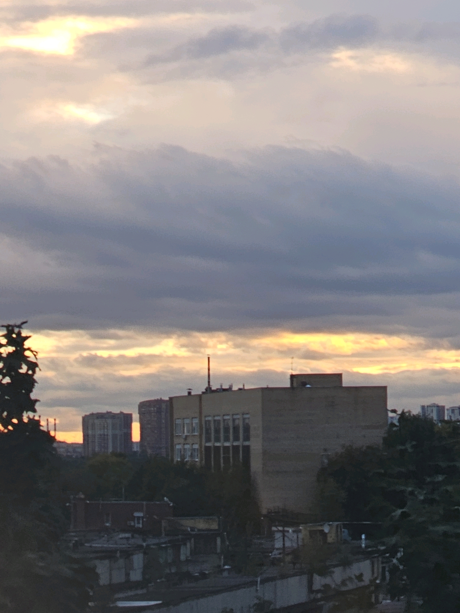 This is a medium-angle, low-light, horizontal photograph taken outdoors in the city of Khimki, Russia. The image captures a view of buildings and trees against a cloudy sky. The sun is setting, casting an orange glow on the clouds. In the foreground, there are several low-rise structures with flat roofs, some appearing to be sheds or garages, with visible corrugated metal and pipes. A dark-leaved tree is visible in the left foreground. In the mid-ground, a light-brown, multi-story building with visible windows is prominent. Behind it, taller residential buildings are visible in the distance. The sky is filled with a mix of dark and light grey clouds, with streaks of orange and yellow from the setting sun. No people or animals are visible in the image. The overall impression is a slightly hazy, urban landscape at dusk.