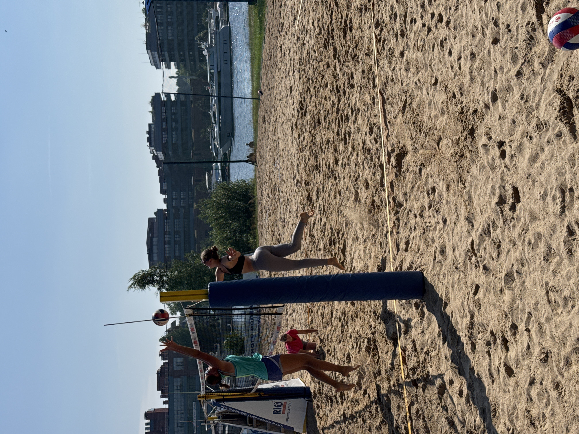 The image depicts a beach volleyball game on a sunny day. Two women are actively playing on a sand court. One woman in a teal tank top and blue shorts is jumping to hit the ball. The other woman, wearing a black sports bra and grey leggings, is also in mid-air, likely in response to the first player's action. A third person in a red shirt is crouched on the sand in the background. The volleyball court is set up with a net and poles. Beyond the court, there is a body of water with several yachts docked. In the background, modern buildings with flat roofs are visible, characteristic of the St. Petersburg cityscape. The overall environment suggests an urban recreational area by the waterfront.
