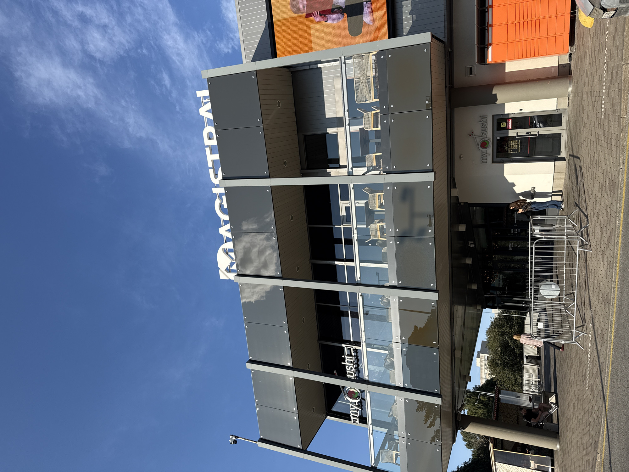The image shows the Magistral shopping center in Tallinn, Estonia, on a sunny day. The building features a modern architectural design with dark grey cladding and glass balconies. On the upper level, a prominent sign reads "MAGISTRAL" in white lettering. Below, the "my sushi" restaurant is visible, with its logo and glass doors. In front of the restaurant, a woman in a dark jacket stands near a metal barrier. To the right of the entrance, a bright orange parcel locker system is installed. In the background, trees and a light-colored building are visible under a clear blue sky. Several people are present on the pedestrian area, including a person sitting on a bench to the left. The scene is captured from a low angle, emphasizing the building's structure and the bright sky.