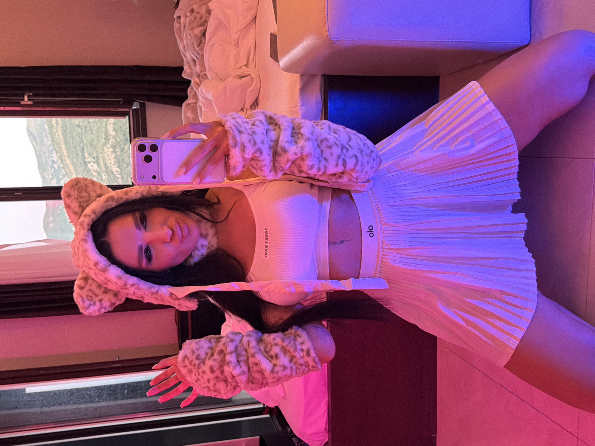 A woman takes a selfie in a room with pink and blue lighting. She wears a fuzzy bear-eared hat, a white Alo-branded sports bra, and a pleated white skirt. A landscape is visible through the window in the background. The location could be Boreti, Montenegro, but this is not discernible from the image.
