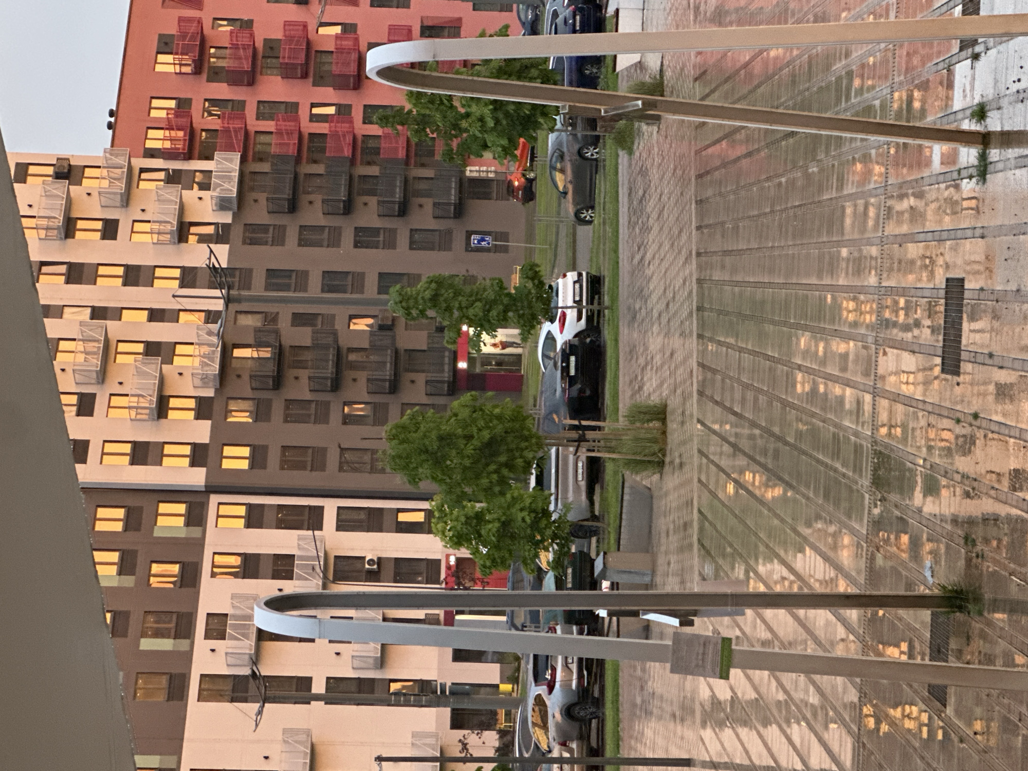 The image captures an urban residential scene in Бараўлянскі сельскі Савет, Belarus, characterized by low-angle warm sunlight or artificial illumination reflected on various surfaces. The foreground displays a wet, paved plaza featuring parallel translucent or reflective panels. Two prominent, light-colored, arching metallic structures rise vertically from this surface, framing the central view. A pole with a small green-edged sign stands behind the left arch.
In the midground, a street runs horizontally, populated by several parked cars including a silver SUV, a white compact car, a dark blue sedan, and a red hatchback. Multiple young trees with green foliage are planted along the street and in landscaping beds. A blue pedestrian crossing sign is visible on a pole. A single person is faintly discernible inside a building entrance with a red frame on the left side of the street.
The background is dominated by multi-story apartment buildings. The building on the left features a facade with alternating light beige and dark brown sections, equipped with numerous balconies presenting white metal railings. The building on the right exhibits a reddish-pink facade with balconies featuring dark red railings. Nearly all visible windows in both buildings reflect a consistent, intense orange-yellow light. The upper portion of the background buildings is partially obscured by an overhead structure or awning extending into the frame. The sky above is a uniform light hue.