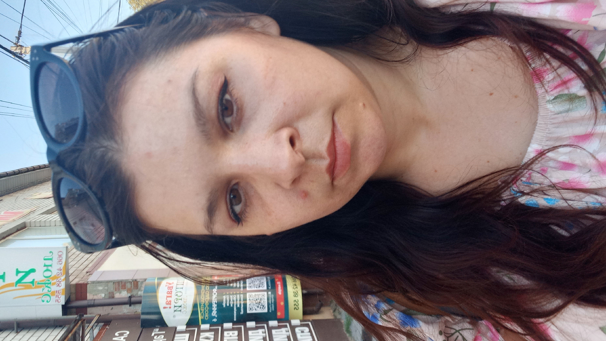 A woman with long, dark hair and sunglasses perched on her head is shown in a close-up, outdoor, selfie-style photograph. She has brown eyes, wears subtle makeup including eyeliner, and has a small blemish on her cheek and another on her chin. Her shirt has a floral pattern with pink, blue, and green elements. In the background, there is a brick building with signage that appears to be in Cyrillic script, suggesting an urban setting. One sign, in particular, displays the letter "N" prominently. The image is taken during daylight hours, indicated by the clear sky and shadows. Based on the distinctive Cyrillic signage and architectural style visible, the location can be identified as Kropyvnytskyi, Ukraine.