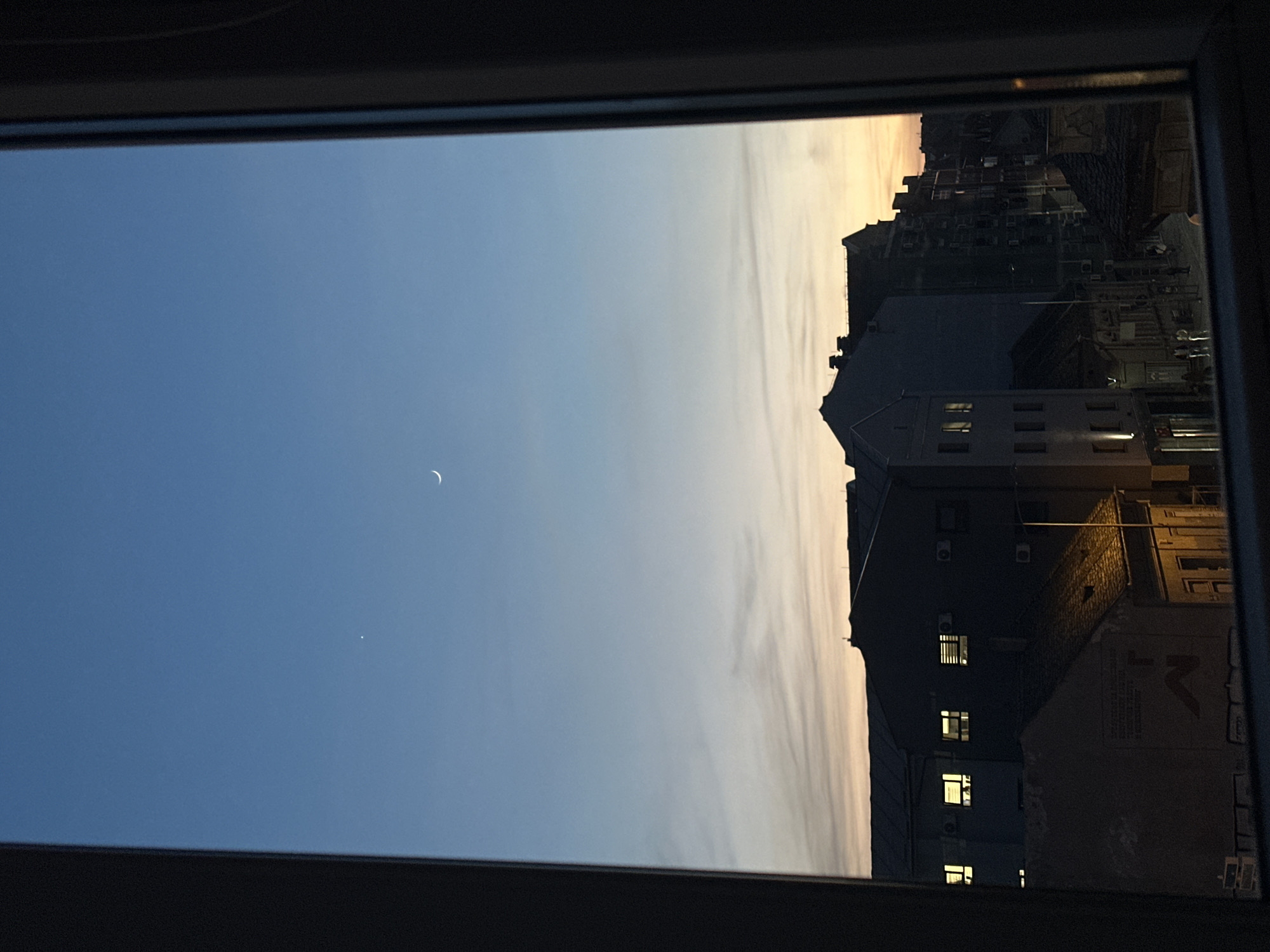 The image shows a view through a window frame of an urban scene in Novi Sad, Serbia at twilight. The sky transitions from a deep blue to a soft yellow-orange at the horizon. A crescent moon and a bright star or planet are visible in the sky. To the right, dark buildings are present with a few lit windows. Details of the buildings include rooftops, windows, and possibly air conditioning units. The scene appears to be taken from inside a building, looking out.