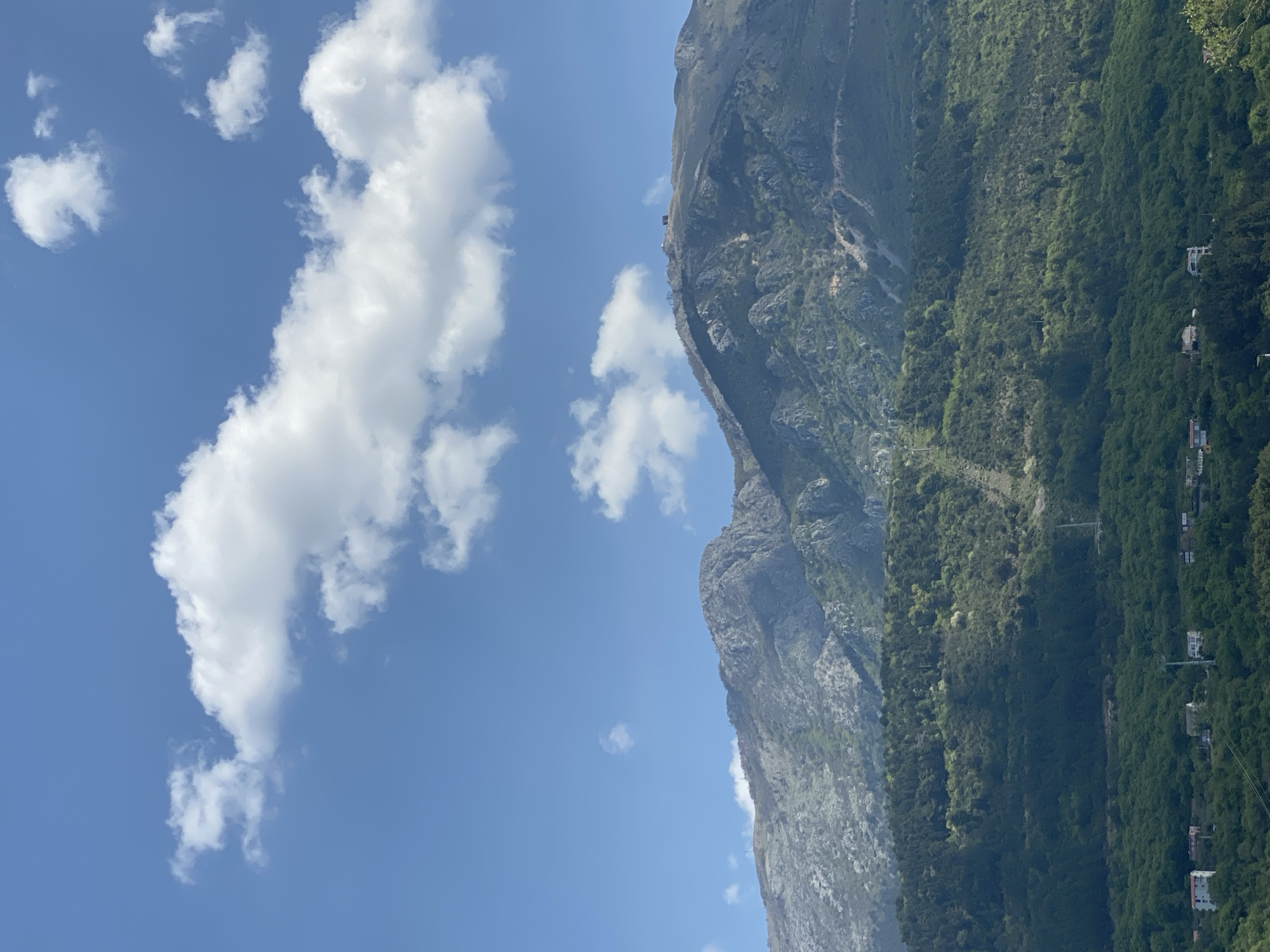 The image captures a vast landscape featuring a towering mountain, lush vegetation, and a clear blue sky with white clouds. The perspective is tilted, making the mountain appear on the right side, rising steeply from the bottom to the top of the frame. The sky occupies the left half and upper portions of the image, displaying a bright, clear blue hue. Scattered across the sky are several fluffy, white cumulus clouds of various sizes.
The mountain face is rugged and rocky, with visible geological contours. Its lower slopes are densely covered in vibrant green foliage, indicating a rich forest. Towards the base of the mountain and extending into the mid-ground, more greenery is visible, interspersed with what appear to be small residential buildings or structures, suggesting a settlement nestled within the natural environment. Thin lines, possibly power lines or cable car cables, traverse the forested area horizontally. A small, dark structure, perhaps a building or a beacon, is barely discernible on the upper ridge of the mountain against the bright sky.
The scene depicts a sunny day, likely late morning or early afternoon, given the bright, direct sunlight and distinct shadows on the mountain. The weather is fair, with a scattering of clouds but no indication of rain. The majestic natural elements combined with the subtle presence of human habitation suggest a location like Kavač, Montenegro, known for its mountainous terrain and picturesque coastal or valley settlements. No visible text is present in the image.