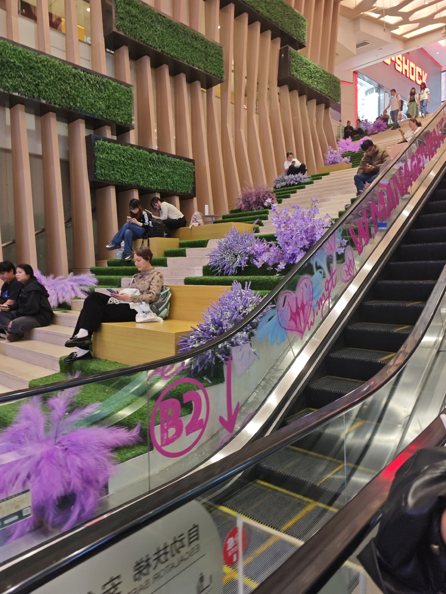 Captured in the Yuzhong District, China, the image depicts a modern interior space. A multi-level staircase, adorned with sections of artificial green grass and purple lavender-like plants, provides seating for several people who are engaging with their mobile phones and reading materials. The staircase is adjacent to an escalator, which has graffiti and other decorative elements on its glass sides, including a pink "B2" symbol with a downward-pointing arrow. The architecture features wooden slats with inset patches of artificial green vegetation above the staircase seating. In the background, a store signage that reads "G-SHOCK" is visible, indicating the presence of retail stores. People can be seen traversing the upper level, in the area that is accessible with the escalator.