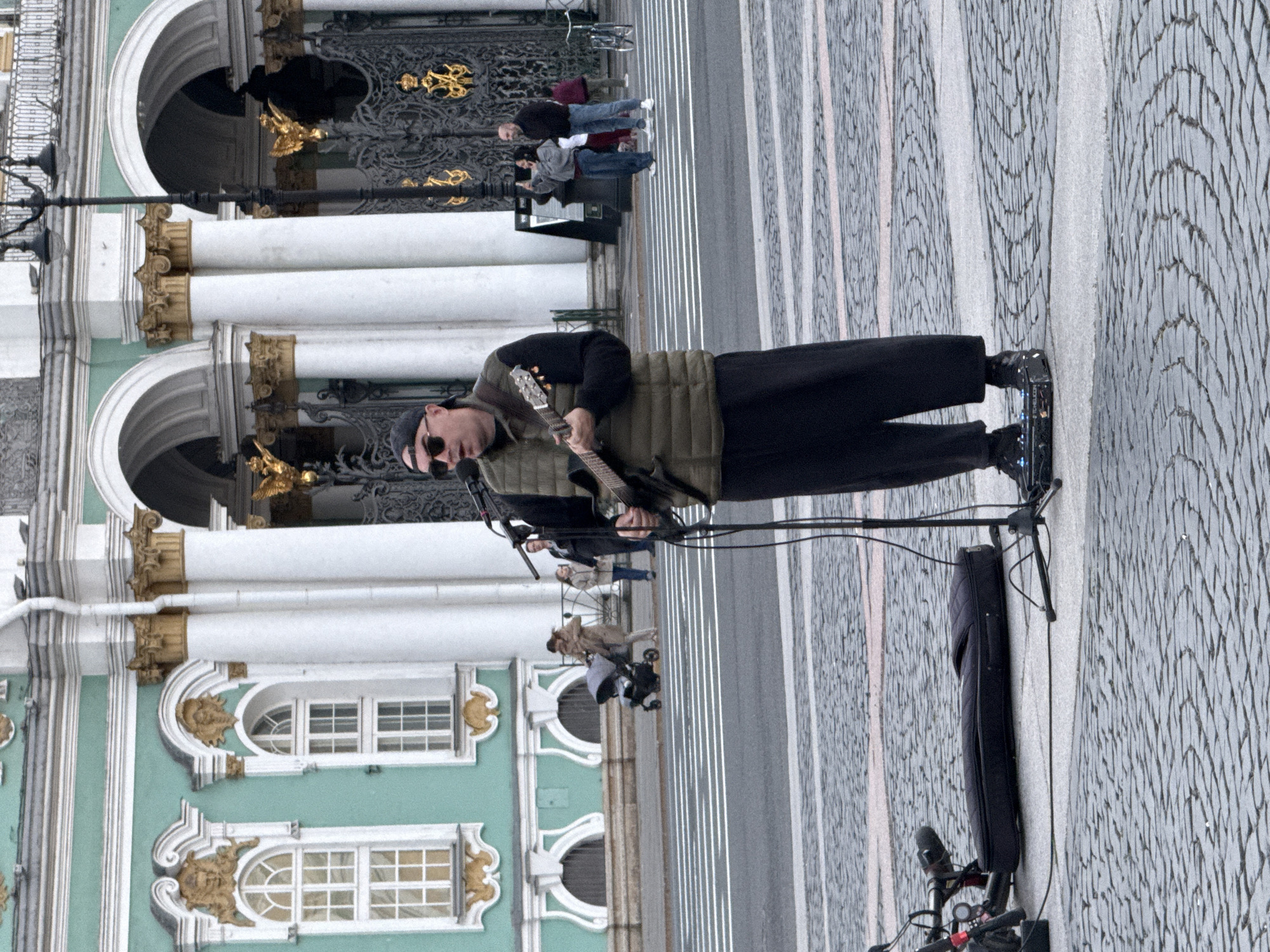 A street musician plays an acoustic guitar in front of a grand, ornate building with pastel green walls and white columns. The musician is wearing sunglasses, a dark t-shirt, a padded vest, and black pants, and is standing next to a black amplifier and microphone setup. The ground is paved with cobblestones and light-colored lines.
In the background, several people are seen walking or observing the musician. One group appears to be a family, with a person holding a baby and another pushing a stroller. Further back, a bicycle is parked. The architecture suggests a historic European city, and the overcast sky indicates it might be a cool, possibly damp day. The scene evokes a sense of urban life and artistic performance in a public space.