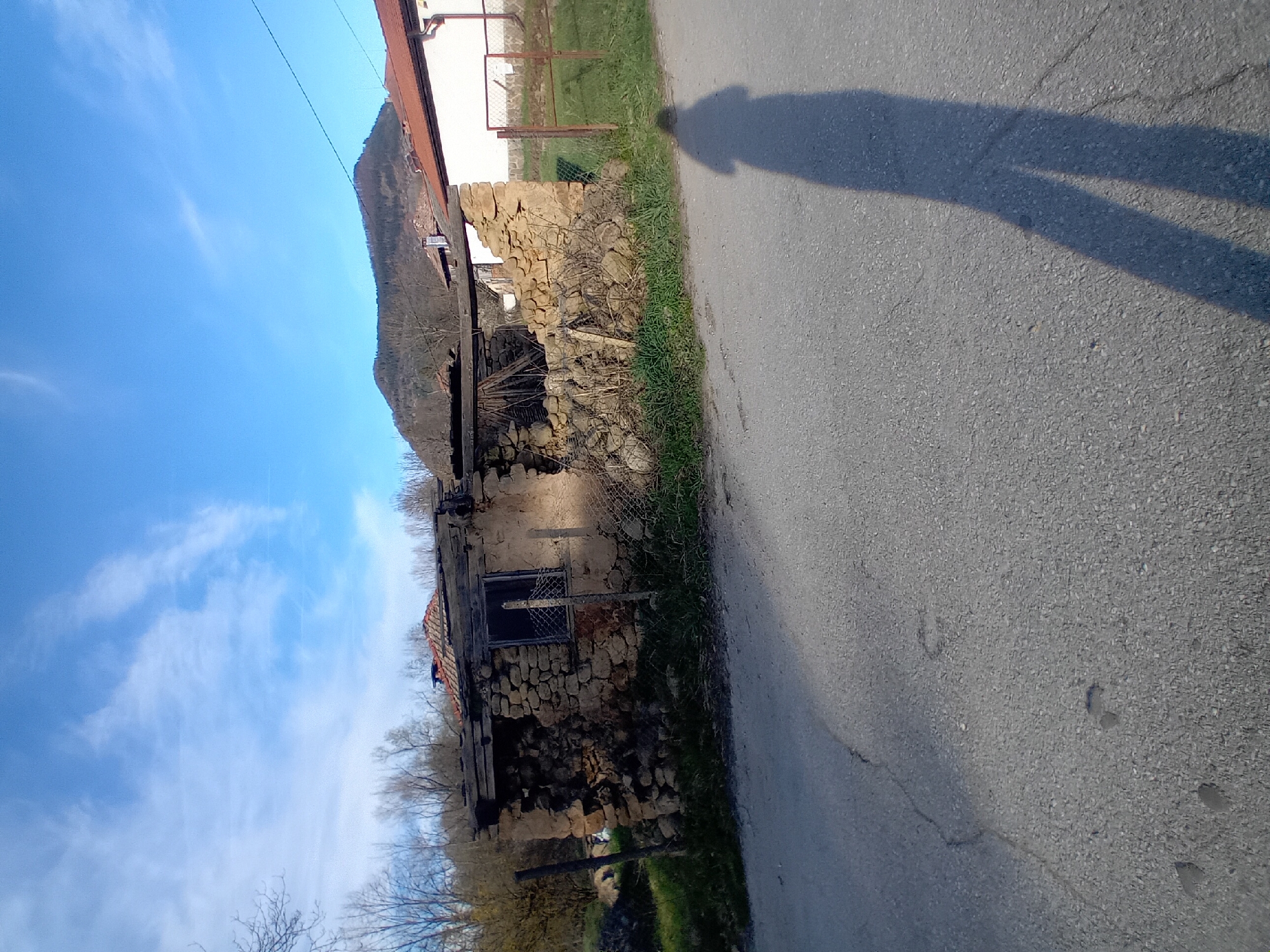 The image shows a weathered stone structure with a partially collapsed roof, possibly an old building or ruin, located adjacent to a paved road. A person's shadow is cast on the road. The environment includes a grassy area between the building and the road. The sky is blue with scattered clouds, suggesting it is daytime. The location is Unknown, Unknown.