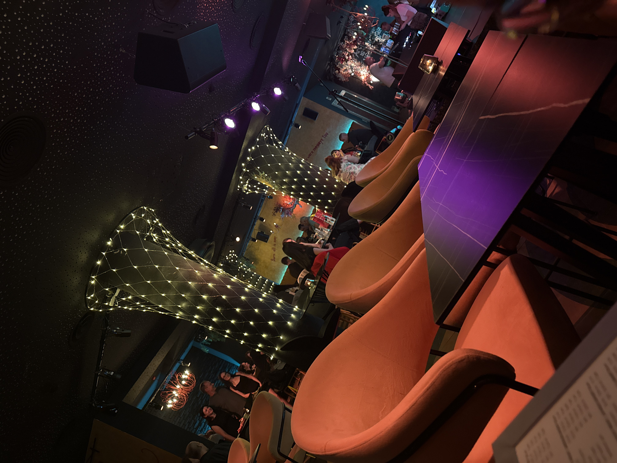 The image displays the interior of a dimly lit establishment, identified as a bar or lounge, located in Sofia, Bulgaria. The perspective is from a low angle, looking across a row of tables towards the back of the venue.
In the foreground, a dark table with a purple-red illuminated, marbled surface extends from left to right. To the left, a portion of a menu is visible on another table. Several high-backed armchairs, upholstered in a vibrant orange-terracotta fabric with dark metal frames, occupy the immediate foreground and midground, positioned around these tables.
Approximately 15-20 individuals are present throughout the scene. In the left-midground, a group of people stands near orange chairs, including a man in a dark shirt, a woman in a black dress smiling, and another woman with glasses. Further back, patrons are seated at various dark tables, engaged in conversations. Towards the right side of the image, a man in a white shirt stands near a microphone stand, suggesting a performance area. A woman in a white top and patterned skirt stands to his right.
The ceiling is dark with a dense, stippled pattern of small white dots, creating a starry effect. Several spotlights and stage lights are mounted on tracks along the ceiling, emitting purple and white light. Two prominent, large, funnel-shaped structures wrapped in warm white fairy lights serve as decorative pillars in the midground, illuminating the space.
Decorative elements on the background walls include a red, ornate chandelier visible behind the left fairy-light pillar. Text is painted on the walls; "We all is one" is visible on the central back wall, and "we always too..." is partially discernible further to the right. On the far right wall, a large, dark speaker hangs from the ceiling, and a floral or tree-like art installation is visible. A single lit candle in a holder is present on one of the dark tables in the midground. The overall lighting scheme is atmospheric, utilizing accent lights and colored illumination.