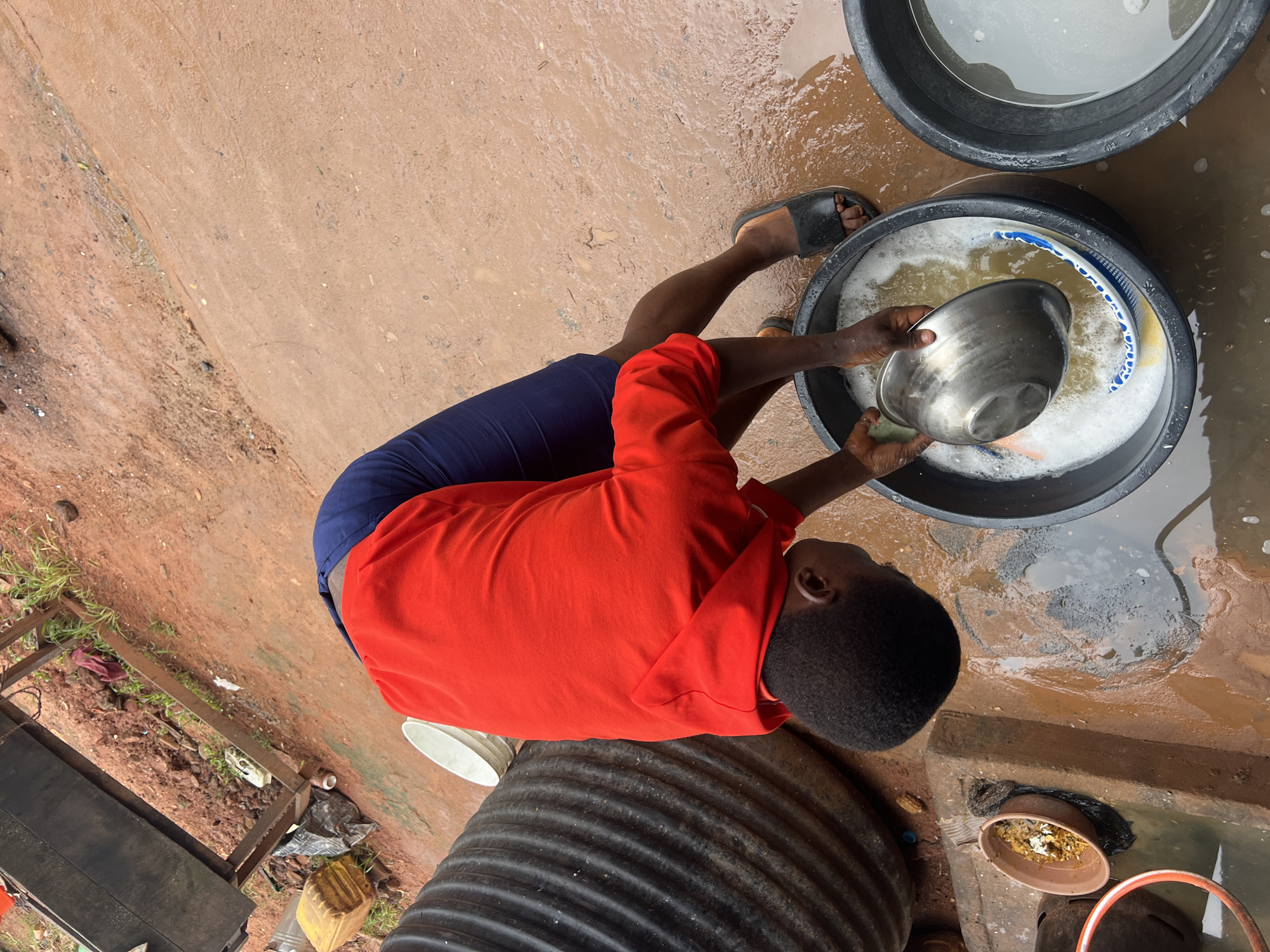 A person wearing a red hooded sweatshirt and dark blue shorts is washing dishes in a large black basin filled with soapy water. They are outdoors in what appears to be a gritty, reddish-brown dirt yard, with evidence of recent rain as the ground is wet and muddy. To the left of the basin, there is a very large, ribbed black container, possibly a water tank. A smaller brown bowl containing food remnants sits on a concrete ledge to the left of the person. In the background, there is a metal structure with a dark surface, and some scattered debris, including plastic containers and a black trash bag. The scene suggests an outdoor domestic or community washing area in Nkpor, Nigeria.