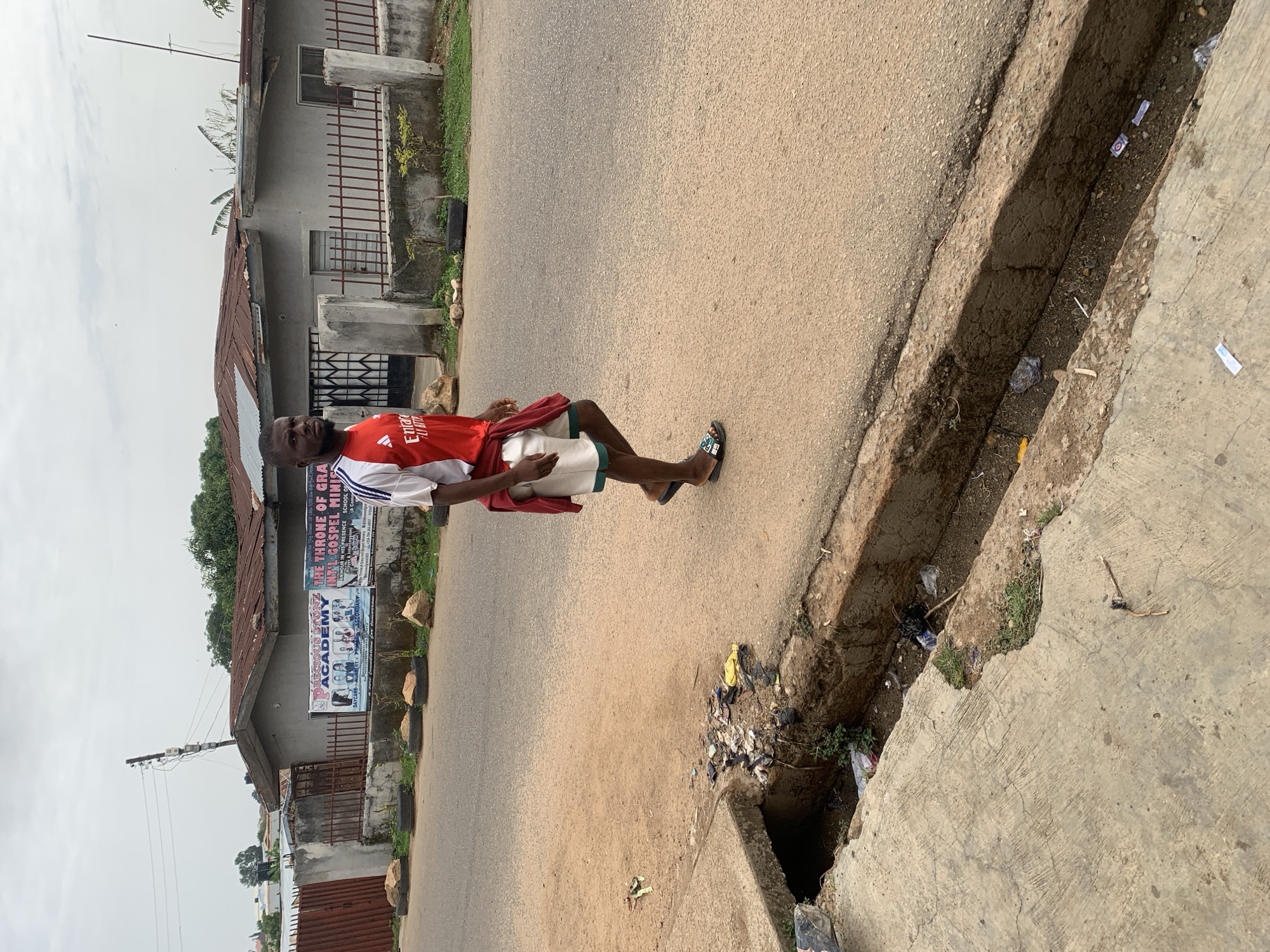 A man stands on a paved road next to a concrete drainage ditch filled with litter in Barakin Akawo, Nigeria. He wears a red and white Arsenal soccer jersey, white shorts with green trim, and black sandals. A building stands behind him with a large sign advertising "PRECIOUS STONE ACADEMY" and "THE THRONE OF GRACE INT'L GOSPEL MINISTRIES." The sky is overcast.