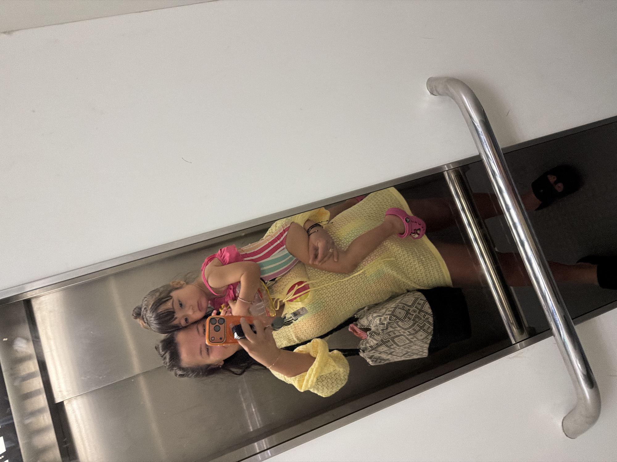 A woman and a young child are reflected in the mirrored elevator doors. The woman is holding the child in her left arm and taking a selfie with her right hand. She is wearing a yellow dress and a patterned bag is slung over her shoulder. The child is wearing a pink and striped swimsuit. The elevator appears to be in a modern building, possibly a hotel or residential complex in Burgas, Bulgaria. The elevator doors are made of brushed stainless steel, and a white wall is visible to the right of the doors. There are chrome handrails on the elevator walls. The floor of the elevator is dark and reflective, showing the reflection of the people and the handrails.