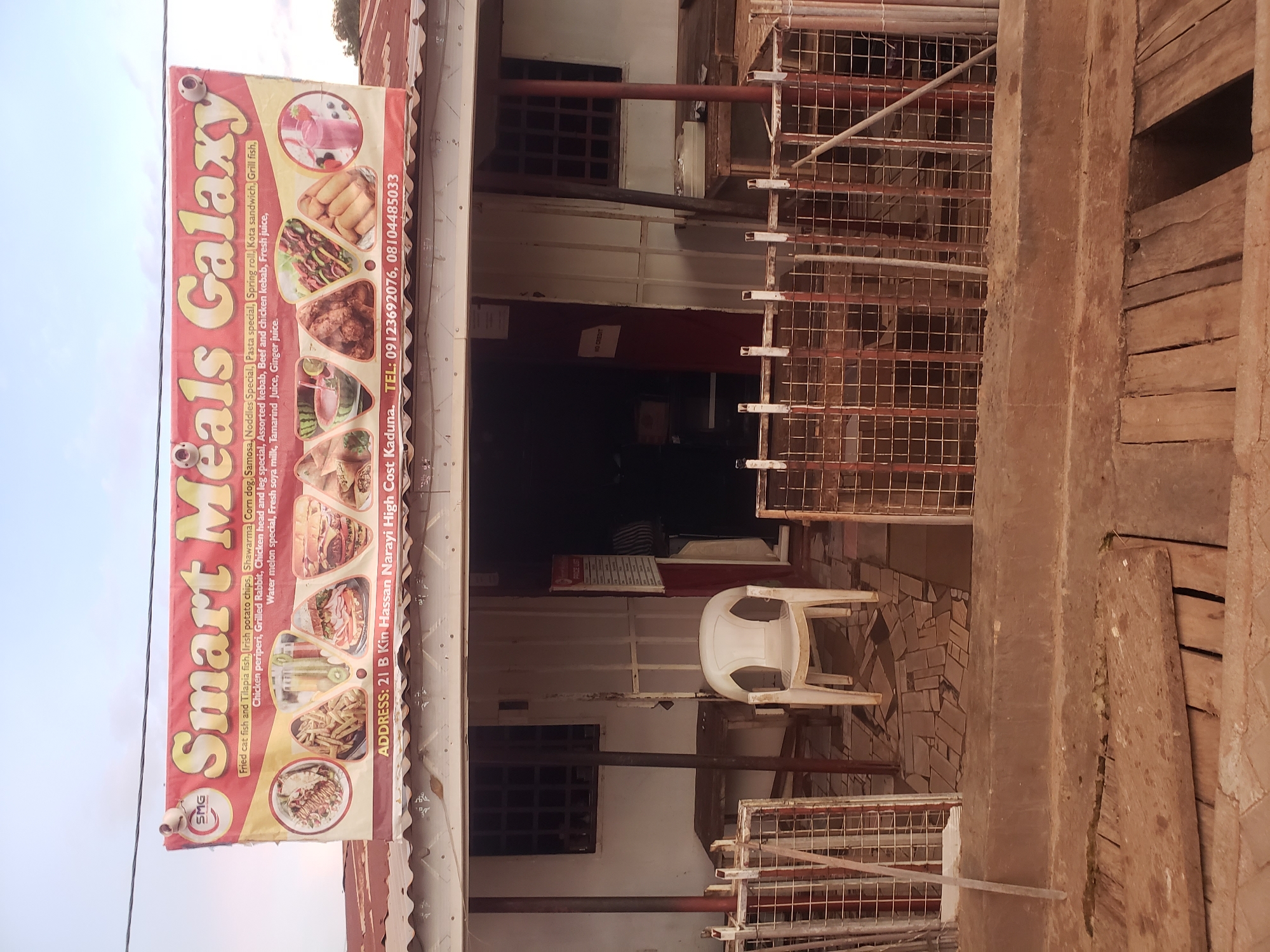 A banner with the text "Smart Meals Galaxy" is displayed above the entrance of a building. The banner lists various food items such as "Fried cat fish and Tilapia fish, Irish potato chips, Shawarma, Corn dog, Samosa, Noodles Special, Pasta special, Spring roll, Kota sandwich, Grill fish, Chicken periperi, Grilled Rabbit, Chicken head and leg special, Water melon special, Fresh soya milk, Tamarind Juice, Ginger juice." It also provides the address "ADDRESS: 21 B Kin Hassan Narayi High Cost Kaduna." and phone numbers "TEL: 09123692076, 08104485033." The entrance area features a white plastic chair and a tiled floor. To the left and right of the entrance are fenced-off areas. The building appears to be a commercial establishment, possibly a restaurant or food stall, located in Narayi, Nigeria, as indicated by the address on the banner.