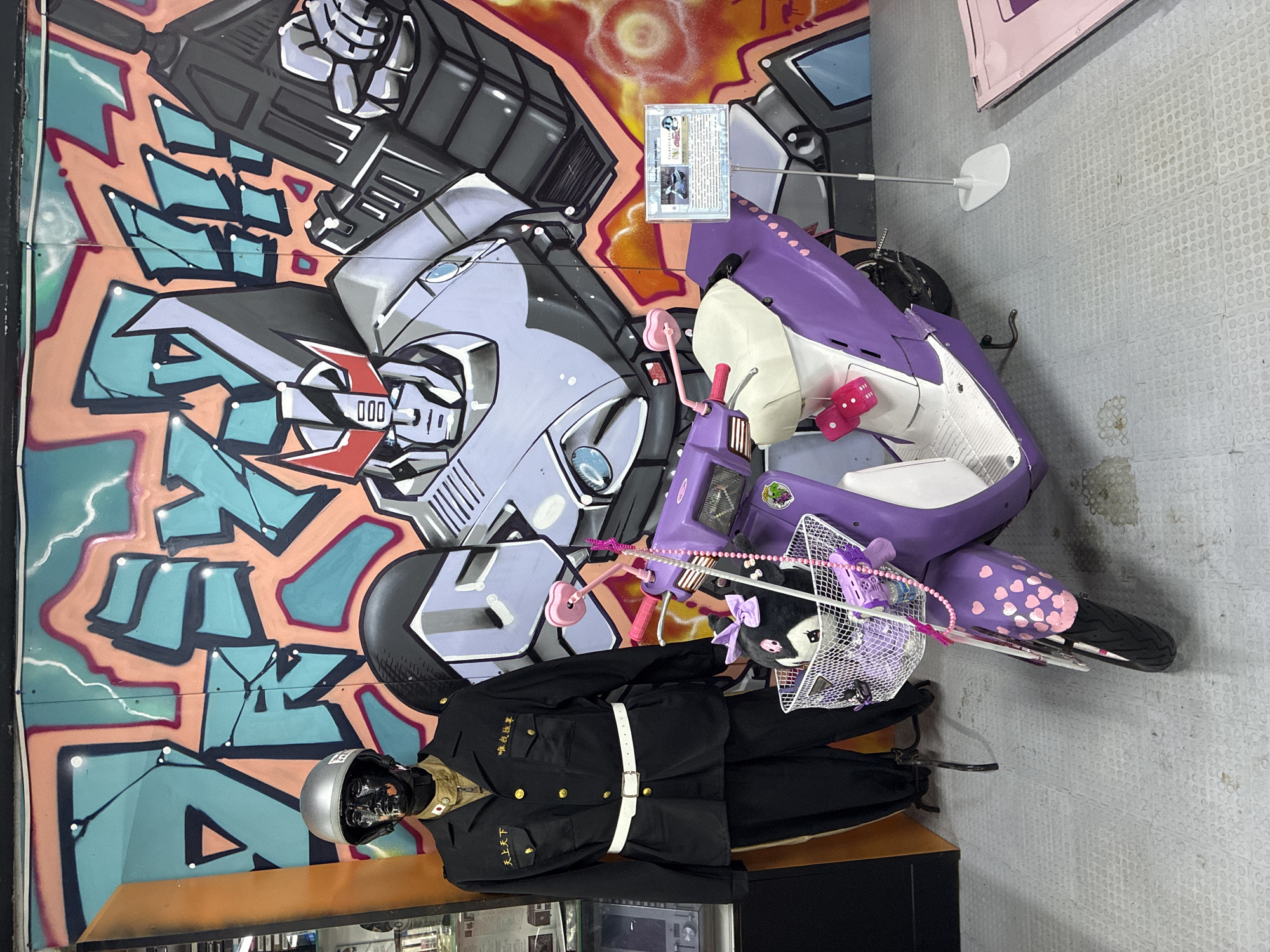 The image shows an indoor display with a graffiti wall featuring cartoonish robot figures in gray and peach tones, with turquoise accents. In front of the wall, a purple and white scooter with pink heart decorations is displayed. To the left of the scooter, a mannequin wearing a black police uniform with gold buttons and a silver helmet is placed on a wooden platform. A white basket filled with toys is attached to the scooter. Above the scooter, a framed card is affixed to the wall. The floor is tiled and the general environment is a well-lit indoor setting. The scene appears to be part of an exhibit or display. There is no indication of the location being in Moscow, Russia.