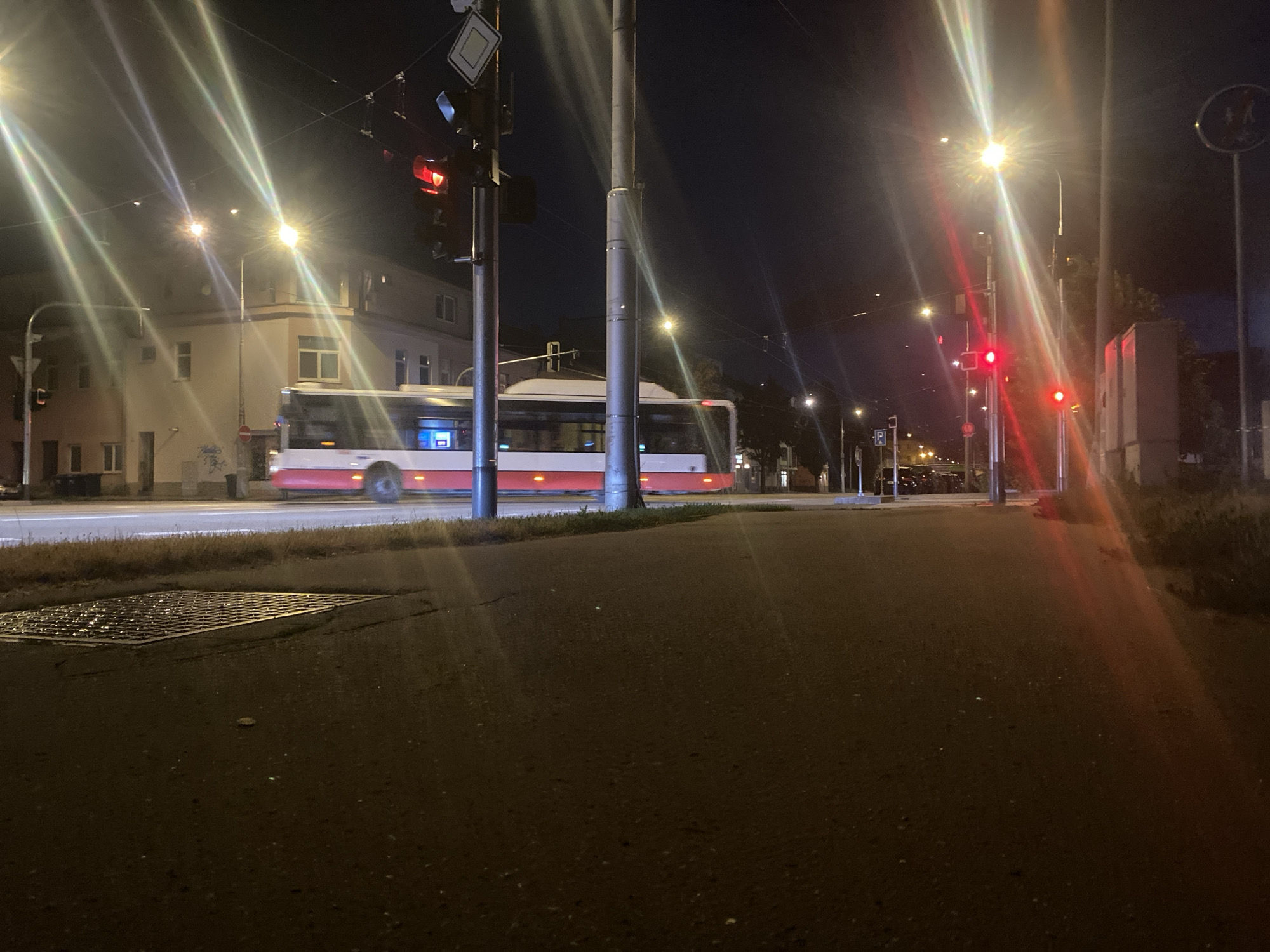 The image depicts a night scene in Brno, Czechia, near a street with a tram. A modern, red and white tram is in motion, its headlights and interior lights illuminating the scene. Traffic lights are visible, with one showing a red signal, suggesting the tram may be stopped or slowing down at an intersection. The street is paved, and a metal grate is present on the sidewalk. Buildings line the street, and streetlights cast bright beams, creating lens flare effects. The sky is dark, indicating it is nighttime. The scene is characteristic of urban public transportation infrastructure at night.