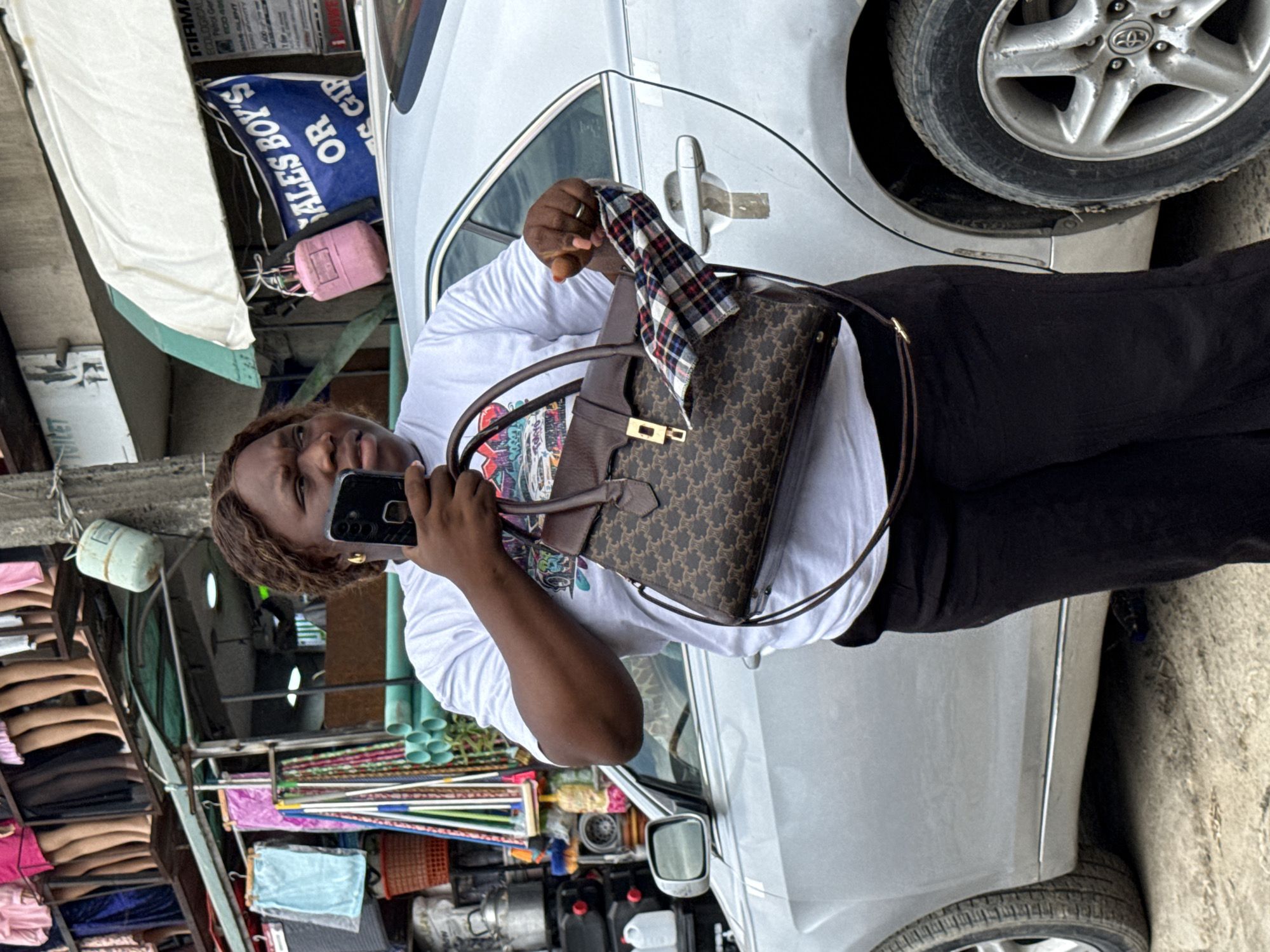 A woman stands in front of a silver sedan, holding a designer purse and a mobile phone. She wears a white t-shirt and black pants. The car is parked on a street in Lagos, Nigeria. Behind her is a vendor's stall displaying various goods, including textiles, pipes, and mannequins. A sign with blue lettering is visible above the stall. The scene depicts a typical street setting in an urban African environment.