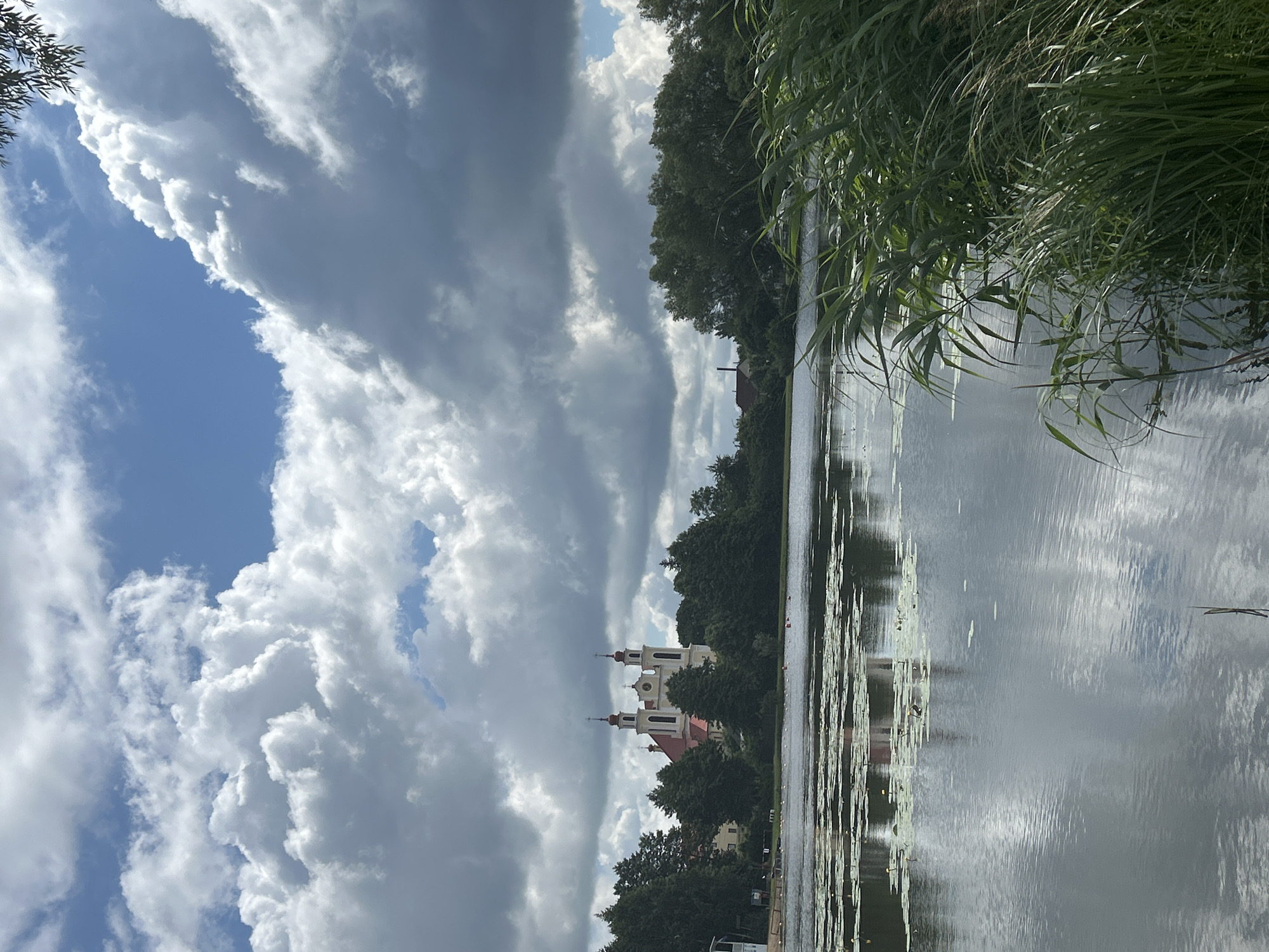 The image depicts a scenic view of a lake in Iwye, Belarus, with a prominent church situated on the far bank. The sky above is filled with dramatic, large cumulus clouds, interspersed with patches of blue. The lake's surface is relatively calm, reflecting the sky and the surrounding greenery. Lily pads are visible scattered across the water.
On the far side of the lake, a white church with red roofing and twin spires stands amidst a dense line of trees. A few other buildings, possibly houses, are visible peeking through the trees. The foreground of the image is dominated by tall, green reeds growing along the water's edge. The overall impression is one of natural beauty and tranquility, with the architectural landmark adding a point of interest to the landscape. No people or animals are clearly visible in the frame.