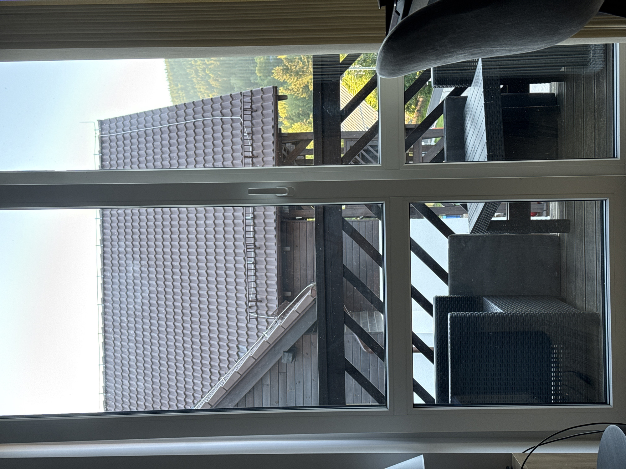 The image shows a view through a window of an outdoor area with a wooden deck and some furniture. On the left side of the image, there is a close-up view of a dark-tiled roof. The scene appears to be from a residential or vacation setting.
In the upper section of the window, we see a portion of a roof with dark, wavy tiles. Beyond the roof, a lush green forest or hillside is visible under a bright, possibly morning or afternoon sky. There are some wooden structures and railings on a deck. In the upper right quadrant, several pieces of dark, woven outdoor furniture are stacked, including what appear to be chairs or lounge seating. The furniture has a wicker-like texture.
The lower section of the window provides a different perspective on the same area. A larger section of a dark-tiled roof is visible, extending horizontally across the frame. A wooden-clad wall of a building is also present, with diagonal wooden beams forming a railing or structural element on a deck. More dark outdoor furniture is visible in the lower right quadrant, arranged on the deck. The furniture is dark and appears to be made of a woven material.
The scene suggests an outdoor living space, possibly a balcony or patio, attached to a building with a distinctive tiled roof. The lighting indicates it is daytime, and the bright sky suggests fair weather. The location context provided, Szklarska Poręba, Poland, places the scene in a mountainous or hilly region known for its natural beauty, which is consistent with the forested background. No people are visible in the image.