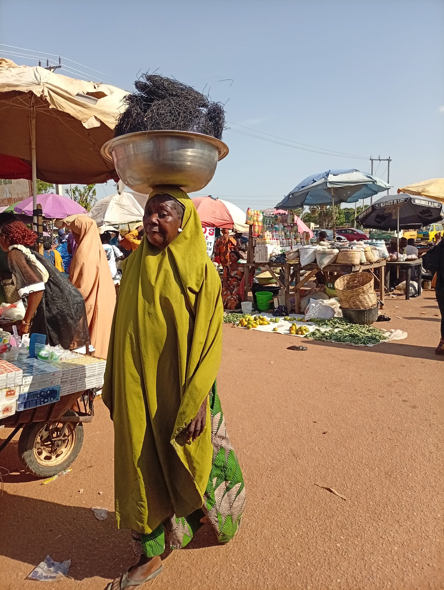 A woman in Kakuri Gwari, Nigeria balances a large metal bowl filled with black material on her head. She wears a green headscarf and robe over a colorful patterned skirt. She stands on a red-brown paved surface. The background shows a bustling market scene with vendors under umbrellas and various goods on display. Other people are visible in the background. A cart laden with merchandise sits to her left.