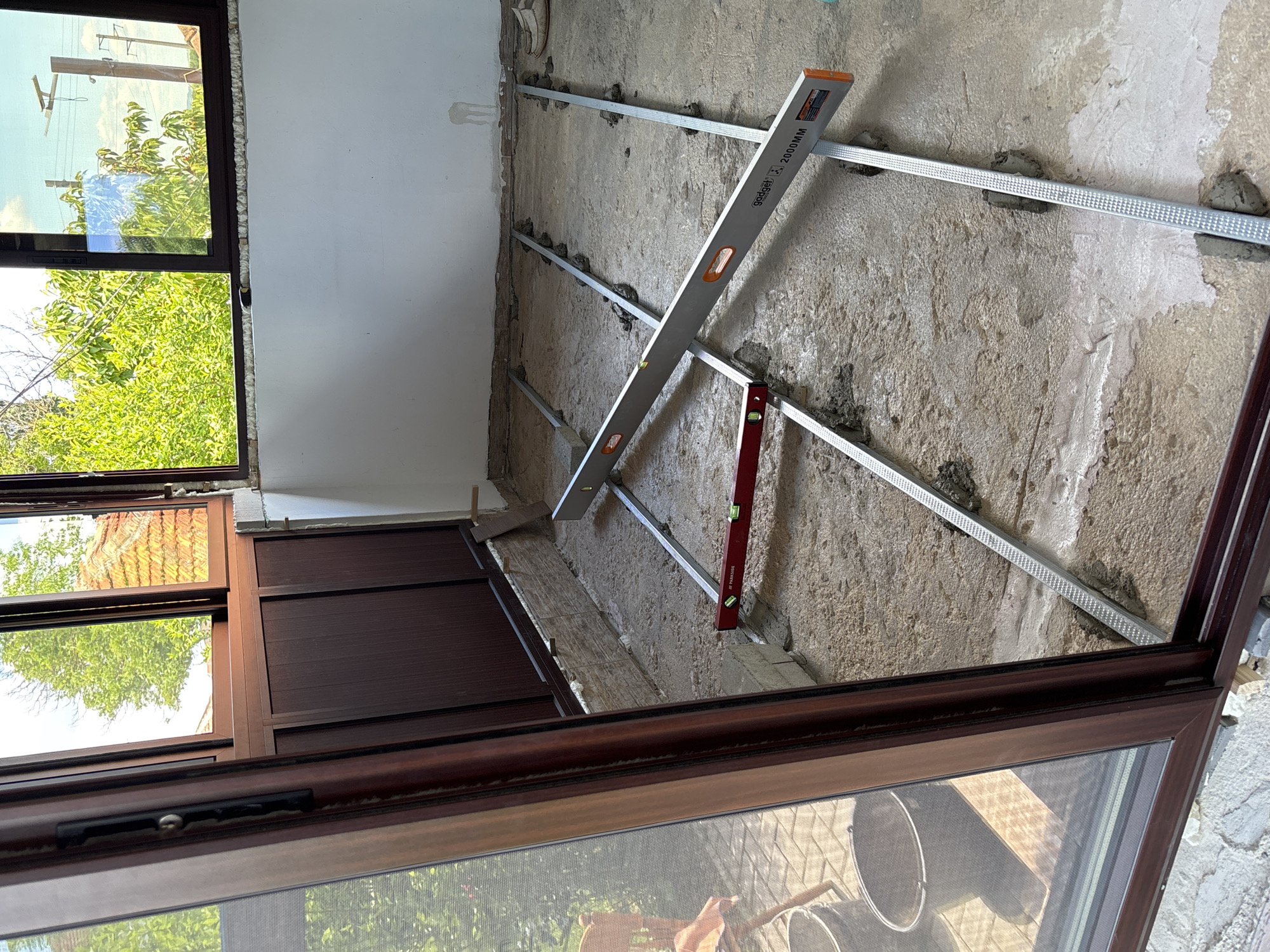 The image depicts an interior construction scene, possibly in a building in Pchelnik, Bulgaria. The focus is on a rough concrete wall being prepared for finishing. Horizontal metal channels are attached to the wall with what appears to be mortar or adhesive. Two levels, a "Gadget" 2000mm and a "Parkside" level, are positioned against the metal channels, likely being used to ensure they are aligned and level. A partially visible brown window frame is in the foreground. Outside the window, green trees and a blue sky can be seen. Inside, the foreground reflection also shows buckets and possibly a hand.