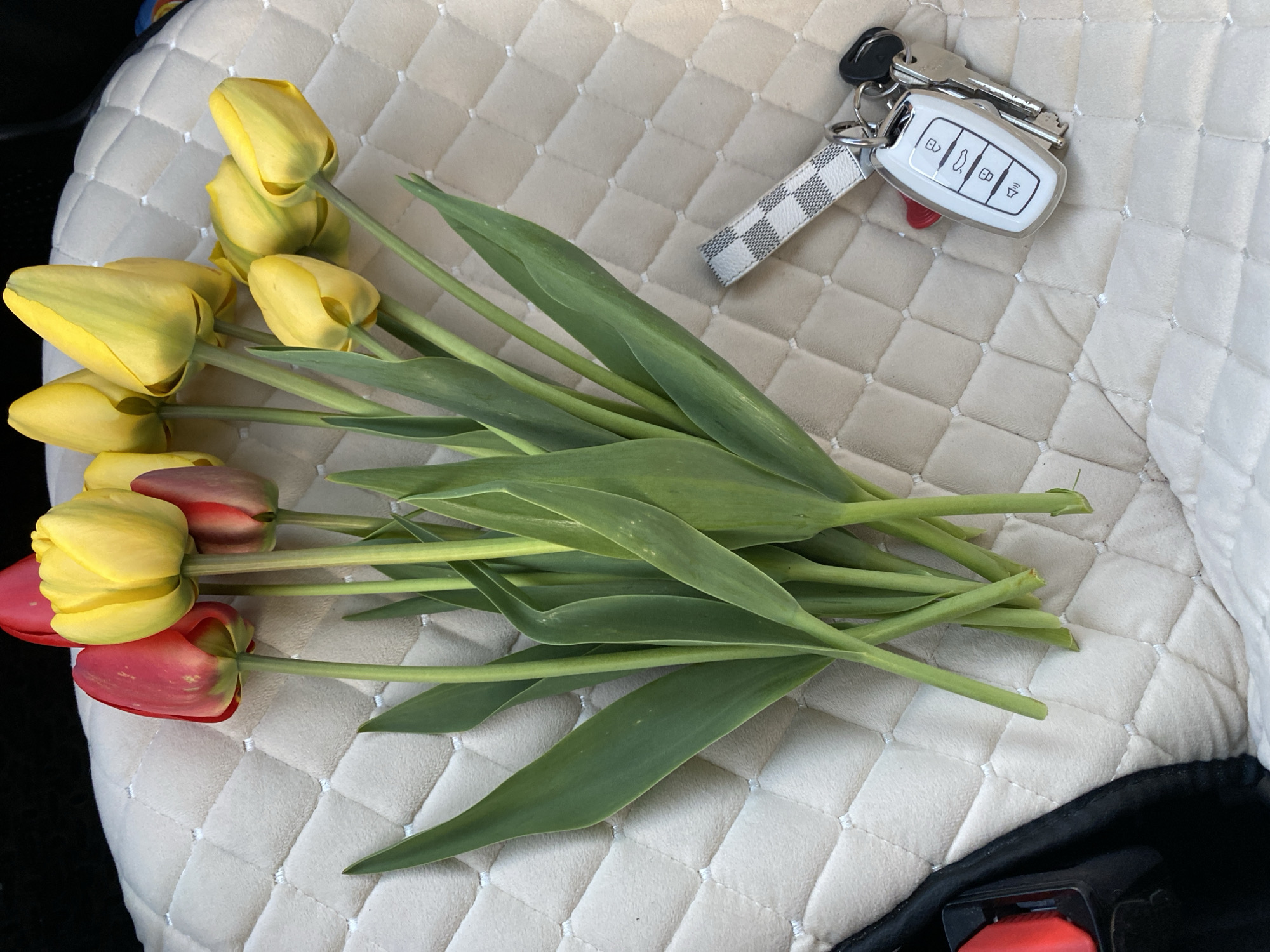 A bouquet of tulips, mostly yellow with some red, lies on a quilted beige surface next to a set of car keys. The keys have a white remote and a white and grey checkered lanyard. Part of a black seatbelt is visible in the bottom right corner. The location in the city of Noviy, Russia is not discernible.