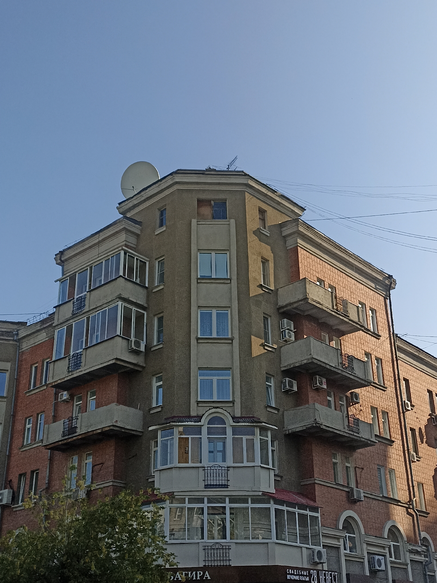 This is a medium-shot, outdoor, eye-level photograph taken on a sunny day, capturing a detailed view of the facade of a multi-story apartment building in Novosibirsk, Russia. The building exhibits a mixture of architectural styles and materials. The left section of the building is constructed with red brick, featuring several balconies with dark metal railings and windows. Above this section, a grey-colored corner protrudes, adorned with multiple glass-enclosed balconies, each with a metal railing on the lowest visible one. This grey section has multiple floors with windows and air conditioning units mounted on the exterior walls. The right side of the building is also primarily red brick, with more balconies that have concrete bases and simple metal railings. This section also features numerous windows and air conditioning units. At the very top of the building, a satellite dish is visible against a clear blue sky. In the foreground, a green tree with dense foliage partially obscures the lower part of the building. The bottom of the image shows a sign with Cyrillic text that appears to advertise a wedding salon. The overall impression is of a typical urban residential building in Russia.