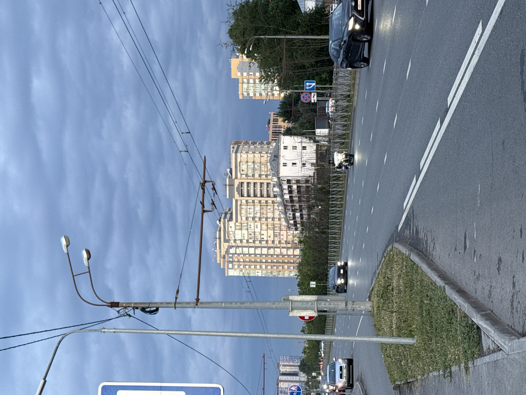 The image depicts a street scene in Kazan, Russia, under a cloudy sky. A multi-lane road stretches into the distance, with cars and a motorcycle in motion. Tall apartment buildings are visible on either side of the road, with some trees and greenery interspersed. A street light pole with two lamps stands prominently in the foreground, along with overhead power lines and trolleybus wires. There are also various road signs, including a pedestrian crossing sign and traffic direction signs. The overall atmosphere suggests an urban environment during either early morning or late evening.