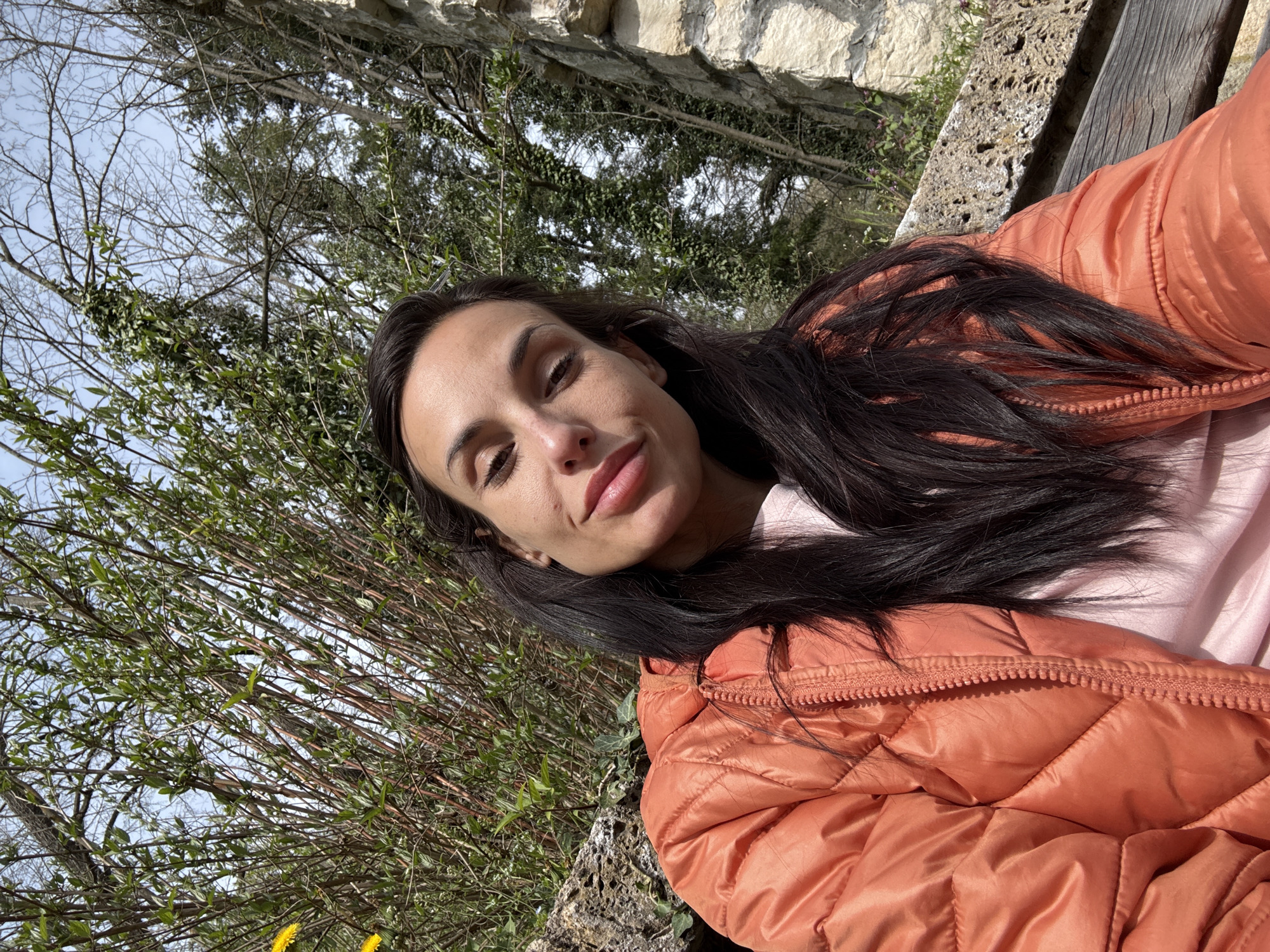 A woman with long, dark hair is lying down outside on what appears to be a wooden bench. She's wearing a light pink top and an orange puffer jacket. Behind her is a stone wall and trees with green foliage and bare branches. The sky is visible through the trees. The location may be in or around Pleven, Bulgaria, given the prompt's specification, but there is no definitive visual confirmation within the image itself.