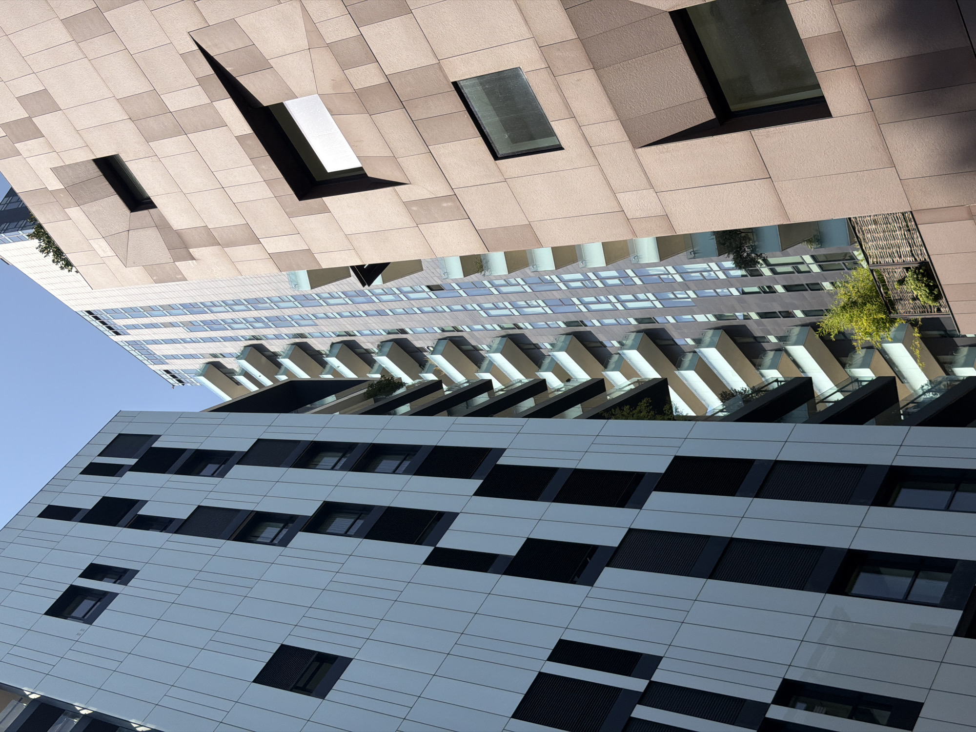 The image captures an exterior view of modern buildings in Milan, Italy, specifically showcasing the Bosco Verticale residential complex. The perspective is a low-angle shot looking upwards, emphasizing the architectural design of the structures.
On the left, a building features a facade composed of light-colored panels with a geometric pattern of dark, recessed rectangular sections, resembling windows or balconies. The verticality of the building is accentuated by the arrangement of these panels and the visible lines of construction.
In the center, a taller, slender skyscraper with a glass facade is visible. Its balconies, edged with white railings and supported by light-colored structures, are tiered and extend outwards, creating a layered effect. Greenery can be seen on some of these balconies.
To the right, a building with a distinct textured facade is prominent. This facade is clad in large, square, beige tiles that are arranged in an irregular, almost pixelated pattern. Some of these tiles are recessed, creating deep shadows and forming angular window openings. One window shows a reflective surface, while another appears darker. A small balcony area with woven fencing and some greenery is visible on this building, nestled between the facade and the central skyscraper.
The sky above is clear and blue, indicating a sunny day. The overall impression is one of contemporary urban architecture, highlighting the interplay of light, shadow, and geometric forms. There are no people or animals visible in the image.