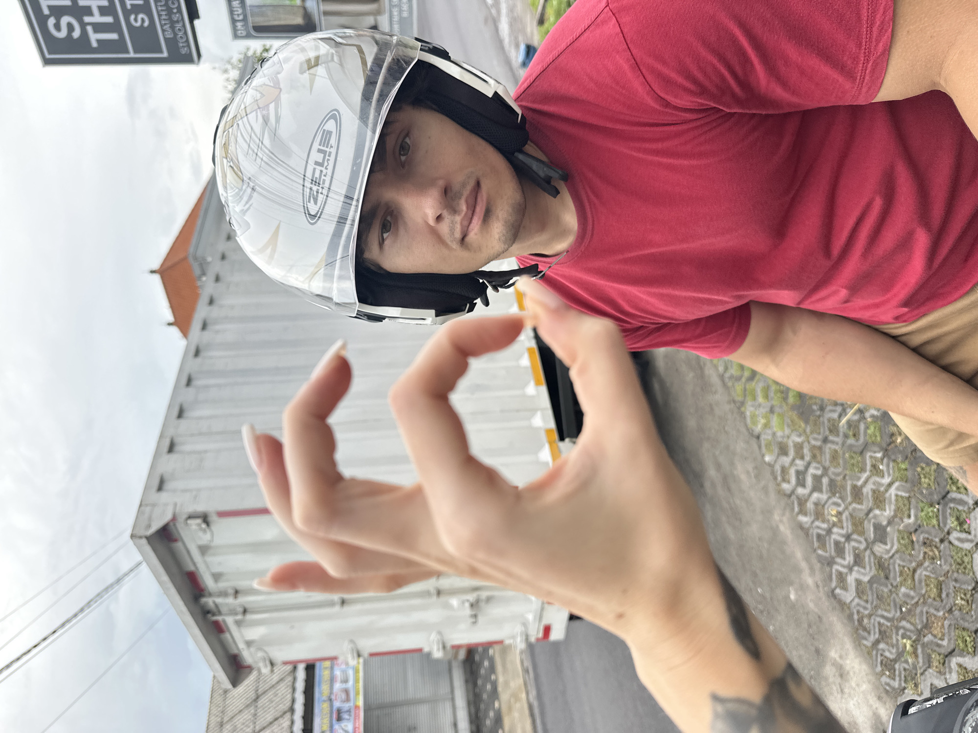 A medium shot captures a man wearing a white helmet and a red t-shirt. He is looking towards the camera, seemingly outdoors. A hand is positioned in the foreground, forming an "OK" gesture. The background includes a white truck, a building with a brown roof, and a street sign that is partially visible with the word "STOOLS." The location is Munggu, Indonesia. The image is oriented vertically.