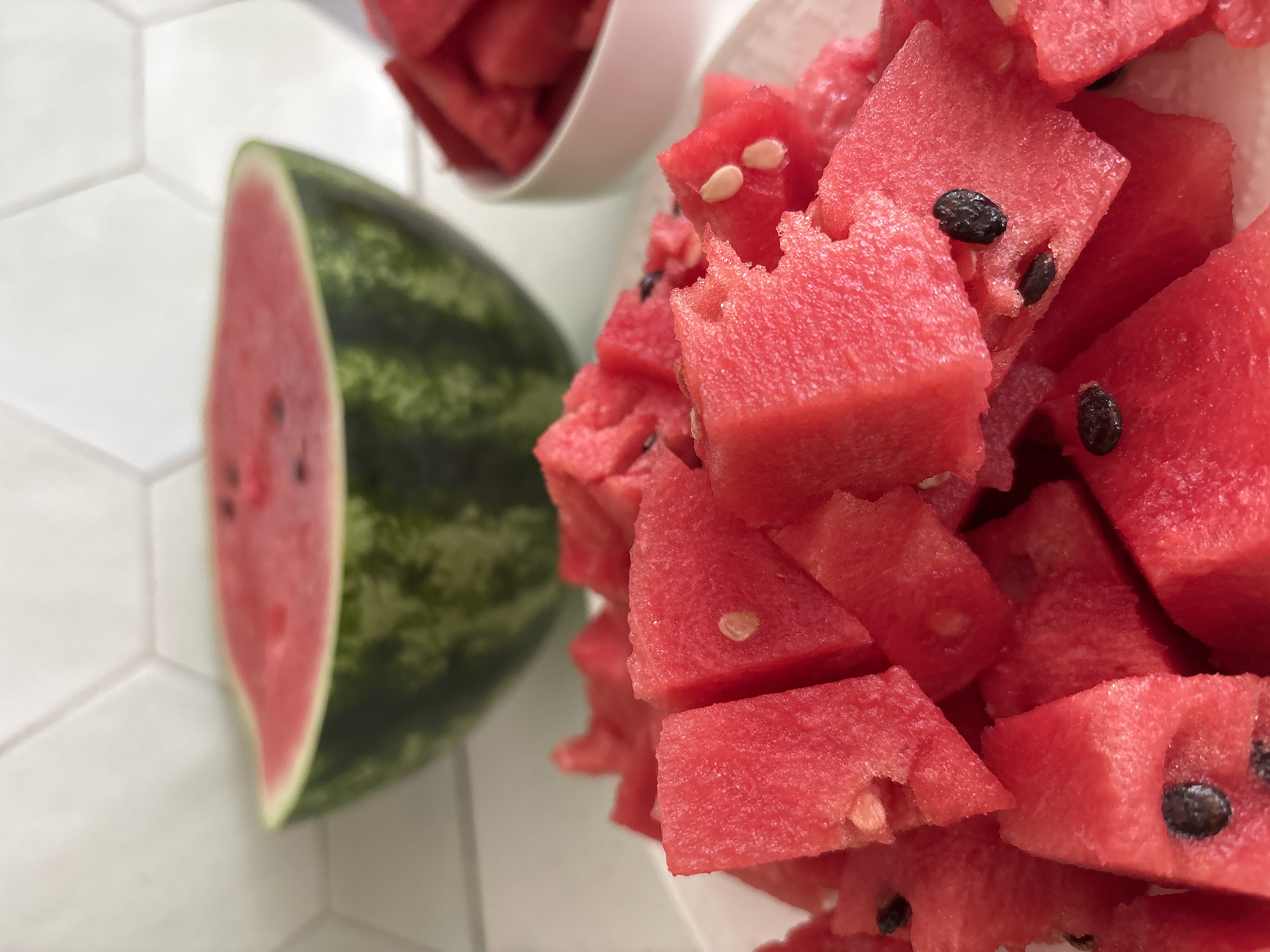 The image displays a close-up view of chopped watermelon pieces, likely prepared for consumption or a refreshing snack. The watermelon is bright red with visible black seeds scattered throughout. Some pieces show slightly jagged edges, indicating they were recently cut. The chopped watermelon is piled into a white container, and some pieces have spilled out. To the left of the chopped watermelon, a large slice of watermelon is visible, showing the green rind and pink flesh. The background consists of a white tiled surface with a hexagonal pattern, suggestive of a kitchen counter or table. The lighting is bright and even, creating a well-lit scene. No people are visible in the image. The overall impression is one of a fresh and inviting presentation of watermelon. The location is noted as Zvenigorod, Russia. There is no visible text in the image.