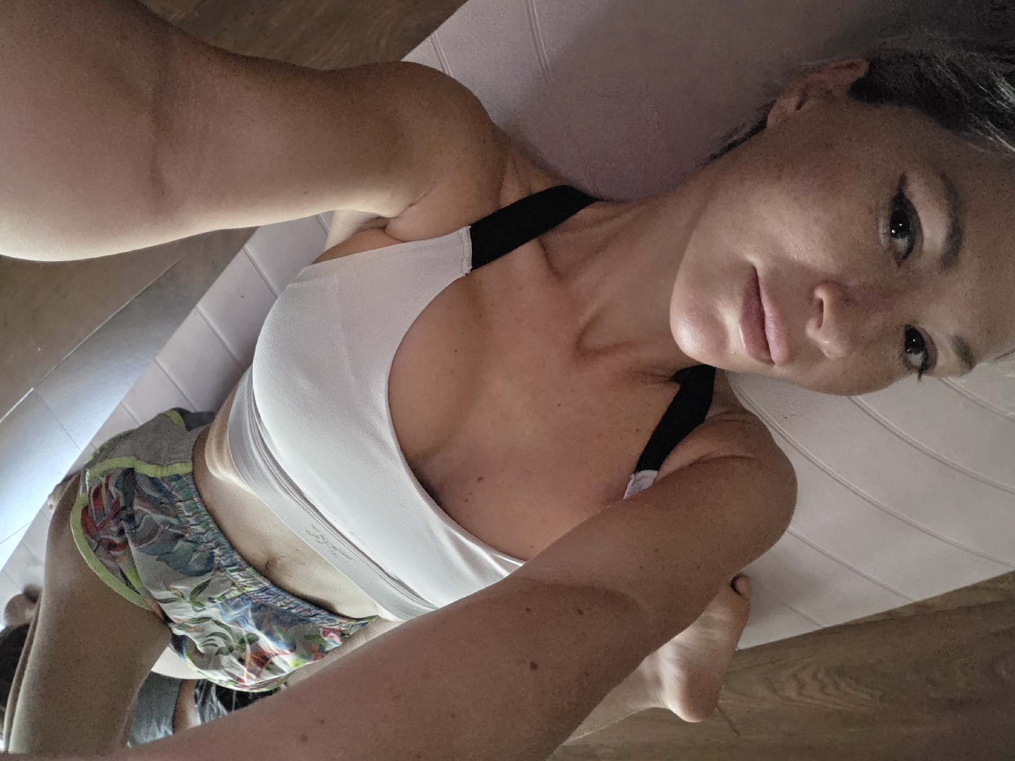 A woman is lying on her back on a white exercise ball, her head is resting on the ball near the bottom of the frame, and her face is turned towards the camera. She is wearing a white sports bra and colorful patterned shorts. Her arms are extended upwards and out to the sides, and her legs are bent with her feet visible at the top of the frame. The background is a wooden floor. The scene is indoors, likely a home gym or studio. The lighting is natural and appears to be coming from the side. The overall impression is one of relaxation and exercise.