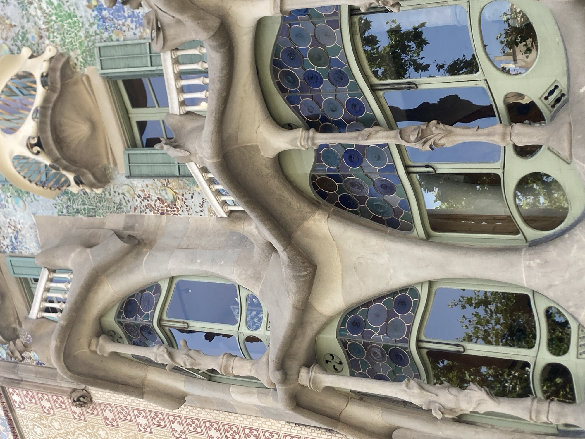 The image showcases the exterior of a building characterized by its unique, organic architectural style. The building's facade features undulating, bone-like stone structures that intertwine with large, irregularly shaped windows. These windows are framed in a pale green material and contain stained glass elements in shades of blue and green, arranged in circular patterns. Through the clear sections of the windows, reflections of trees and a bright blue sky are visible, suggesting it is daytime and the weather is clear. The building's surface is also adorned with colorful mosaic tiles, adding to its distinctive appearance. The overall impression is one of artistic and unconventional design, likely a historic or modern landmark. The image is oriented vertically and appears to be a close-up shot, focusing on the intricate details of the facade. The absence of people and specific events points to a focus on the architectural subject itself. There is no visible text in the image. The style is reminiscent of Antoni Gaudí's work in Barcelona, Spain, known for its nature-inspired forms.