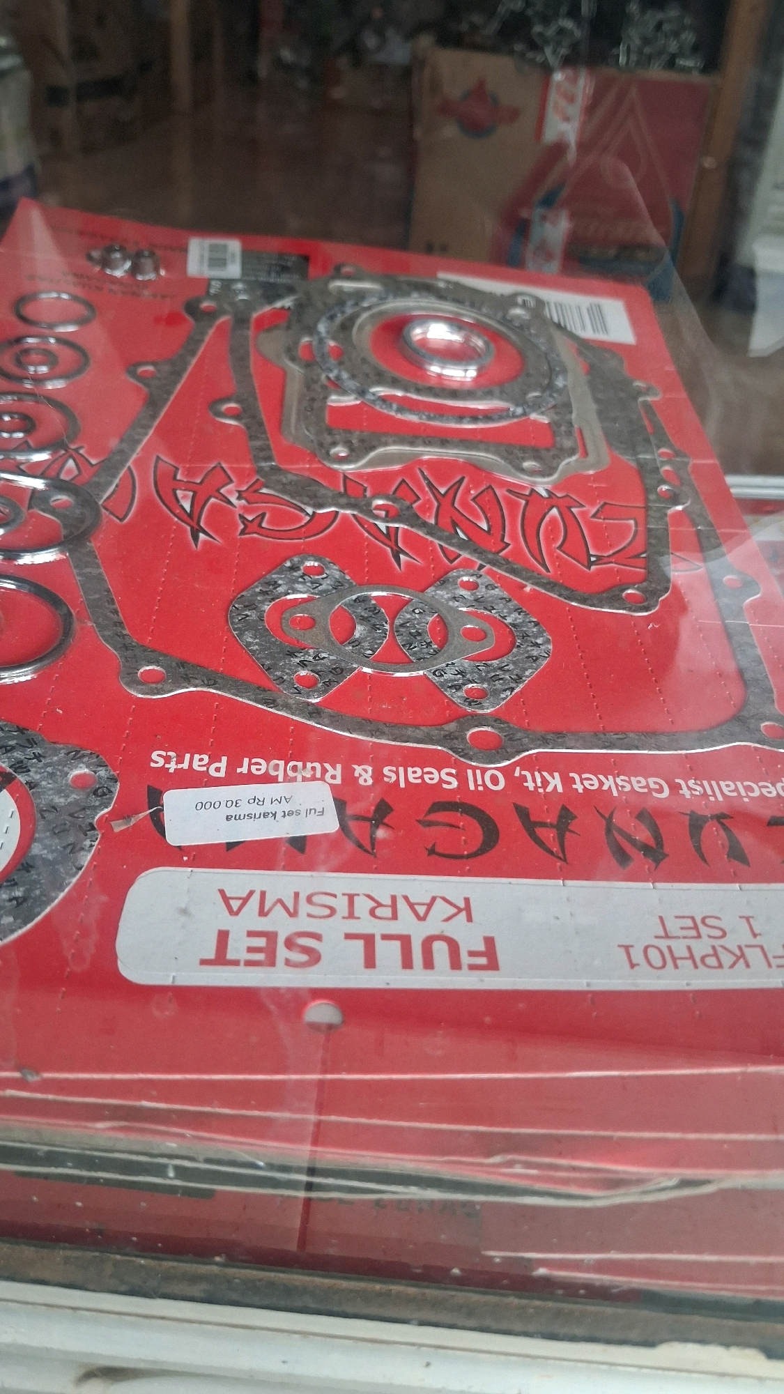 The image displays a close-up, slightly angled view of a red package containing a full set of engine gaskets for a "KARISMA" motorcycle. The gaskets are made of a metallic material with intricate circular and rectangular cutouts, arranged on the red backing. Text on the packaging includes "FULL SET KARISMA", "FUL SET KARISMA AM Rp 30.000", and "Specialist Gasket Kit, Oil Seals & Rubber Parts". The scene is indoors, likely a shop or market stall in Karang Sari, Indonesia, as indicated by the text on the packaging. The packaging is made of plastic and appears to be hanging or displayed. The lighting is moderate, and the focus is on the product.